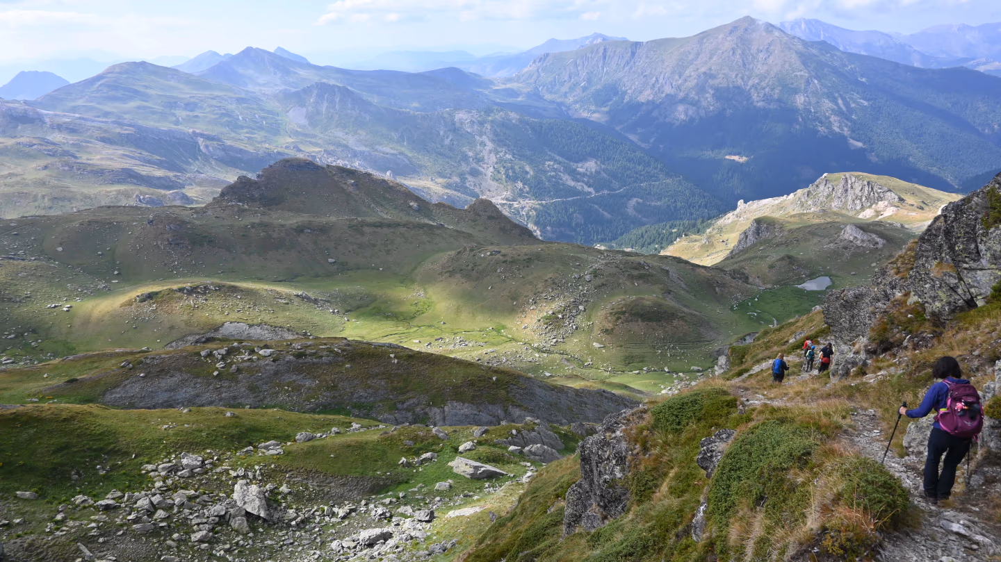 Hikers trekking down a green ridge on the Via Dinarica trail in Kosovo, surrounded by mountain views.