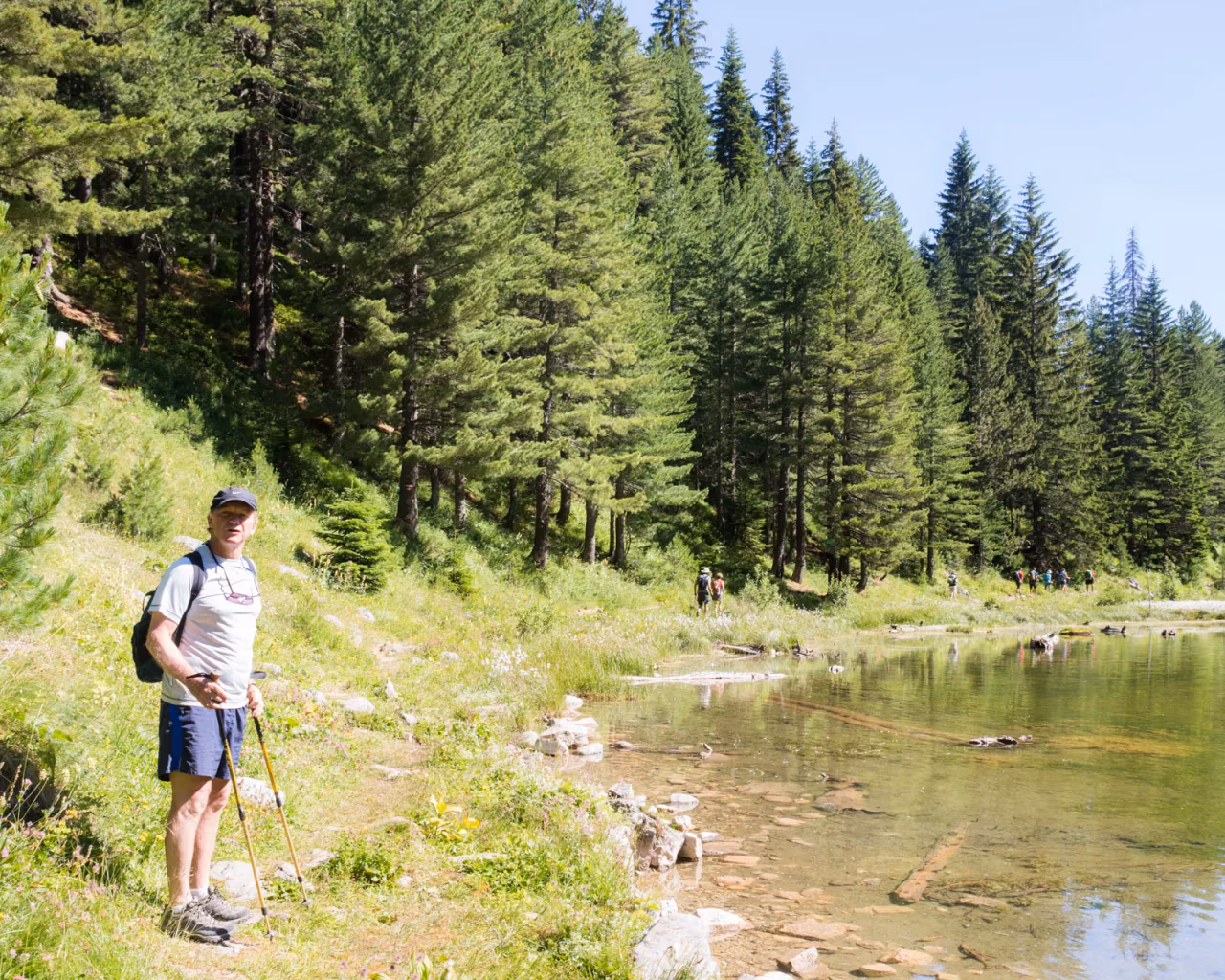 Hiker standing beside a forest lake on Kosovo’s Via Dinarica trail, with clear water and tall pines.