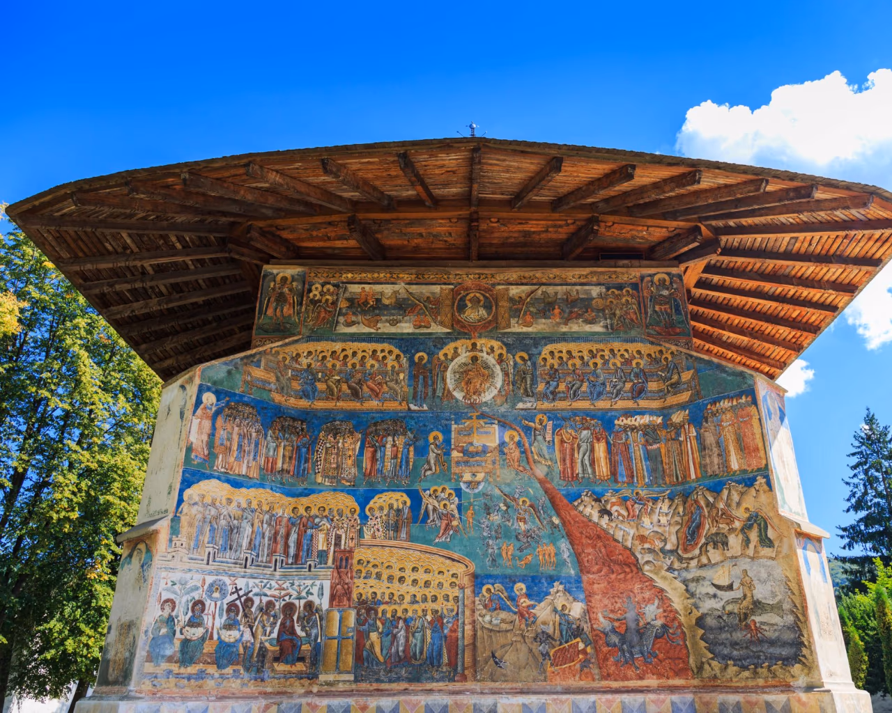 Painted monastery exterior with colorful biblical frescoes under a wooden roof.