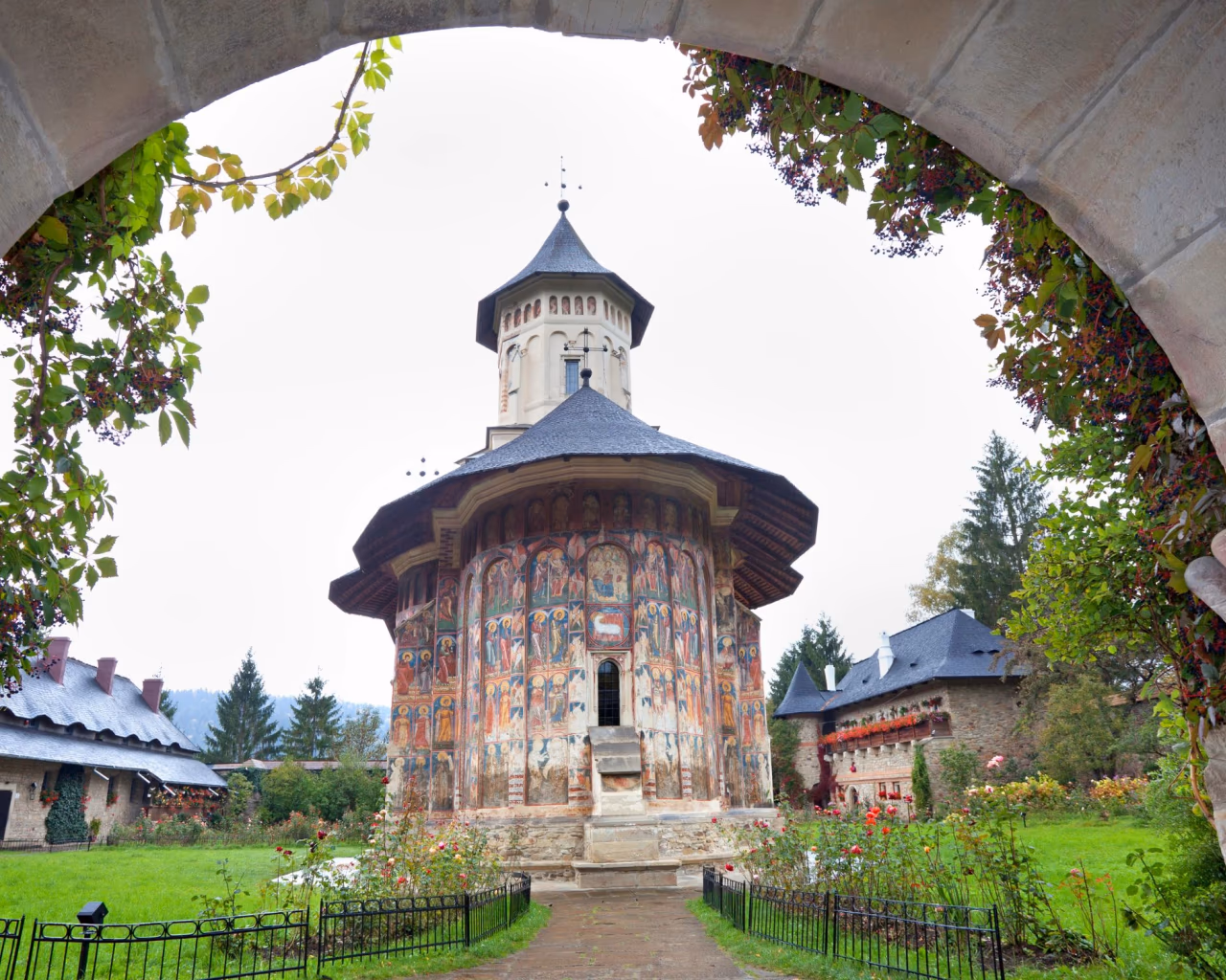 Bucovina monastery framed by a stone arch, with colorful frescoes and lush garden in the courtyard.