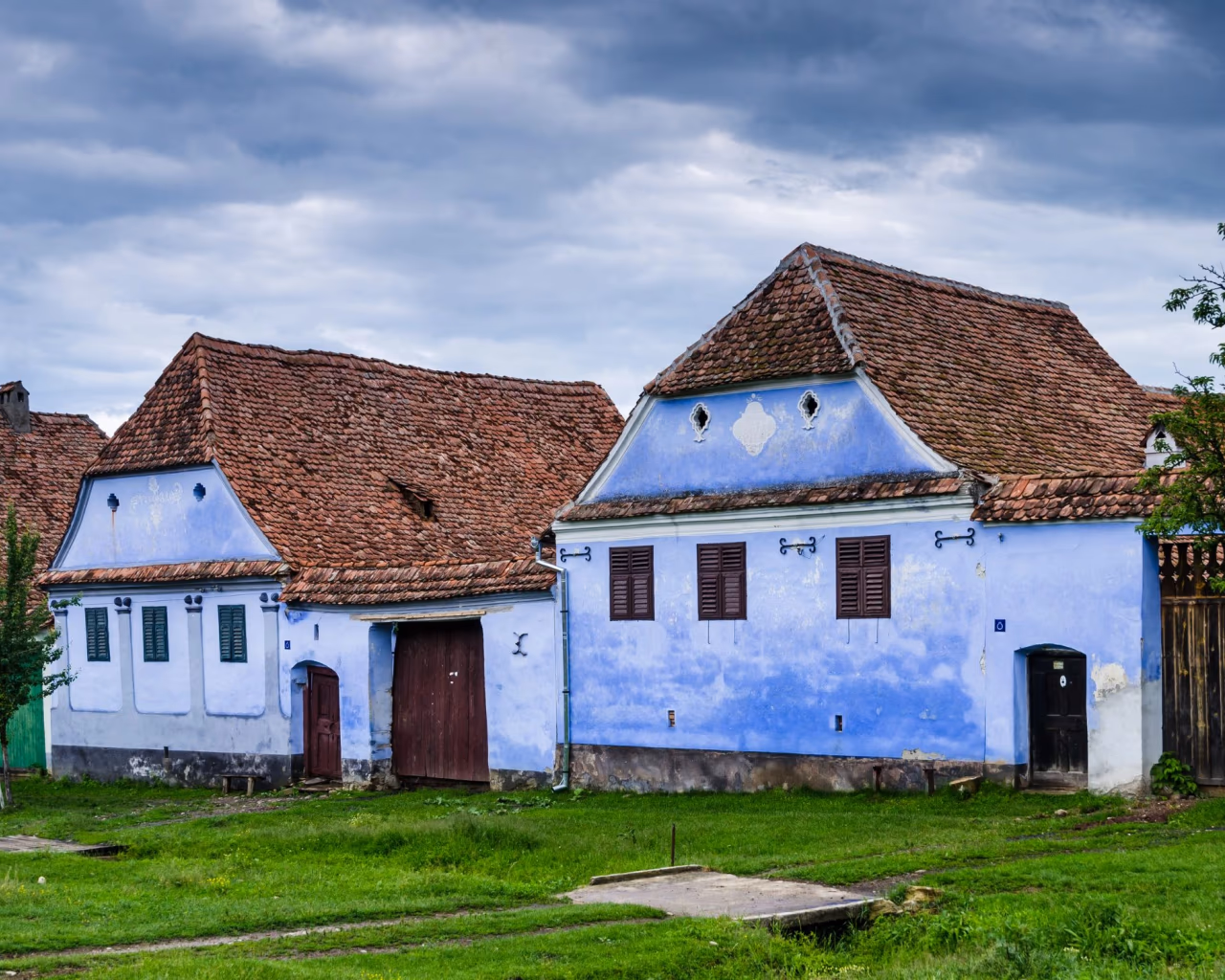 Traditional blue houses with red tiled roofs in a rural Romanian village under a cloudy sky.