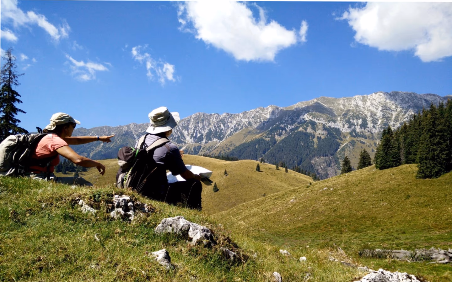 Two hikers rest on a grassy hill, checking a map, with rocky Carpathian peaks and forests stretching before them.