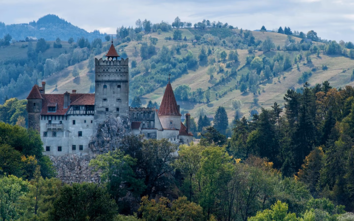 Bran Castle in Transylvania, with red roofs and stone towers, surrounded by green forests and rolling hills.