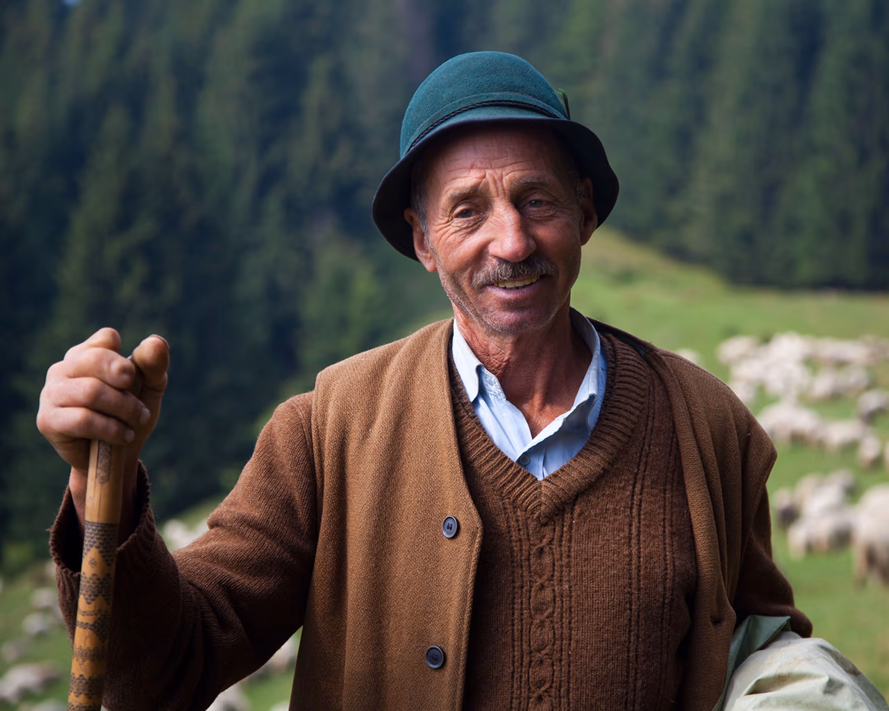Smiling shepherd in a wool coat and green hat holds a wooden staff, with his flock grazing behind him in a mountain field.