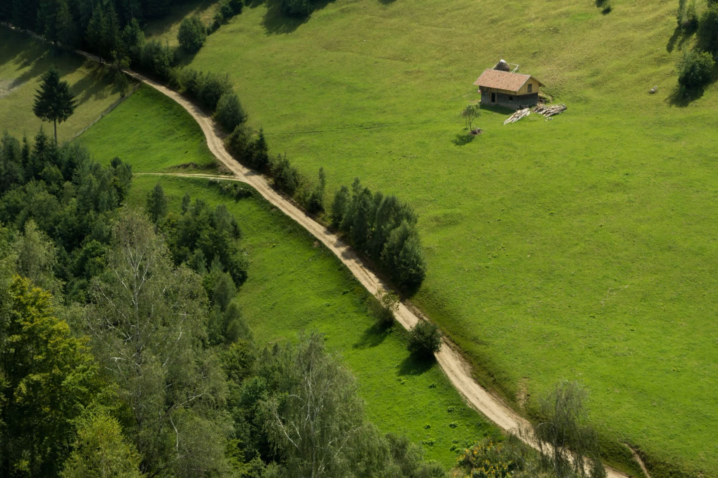 Isolated wooden cabin on a green hillside, reached by a dirt path, surrounded by open fields and scattered trees.