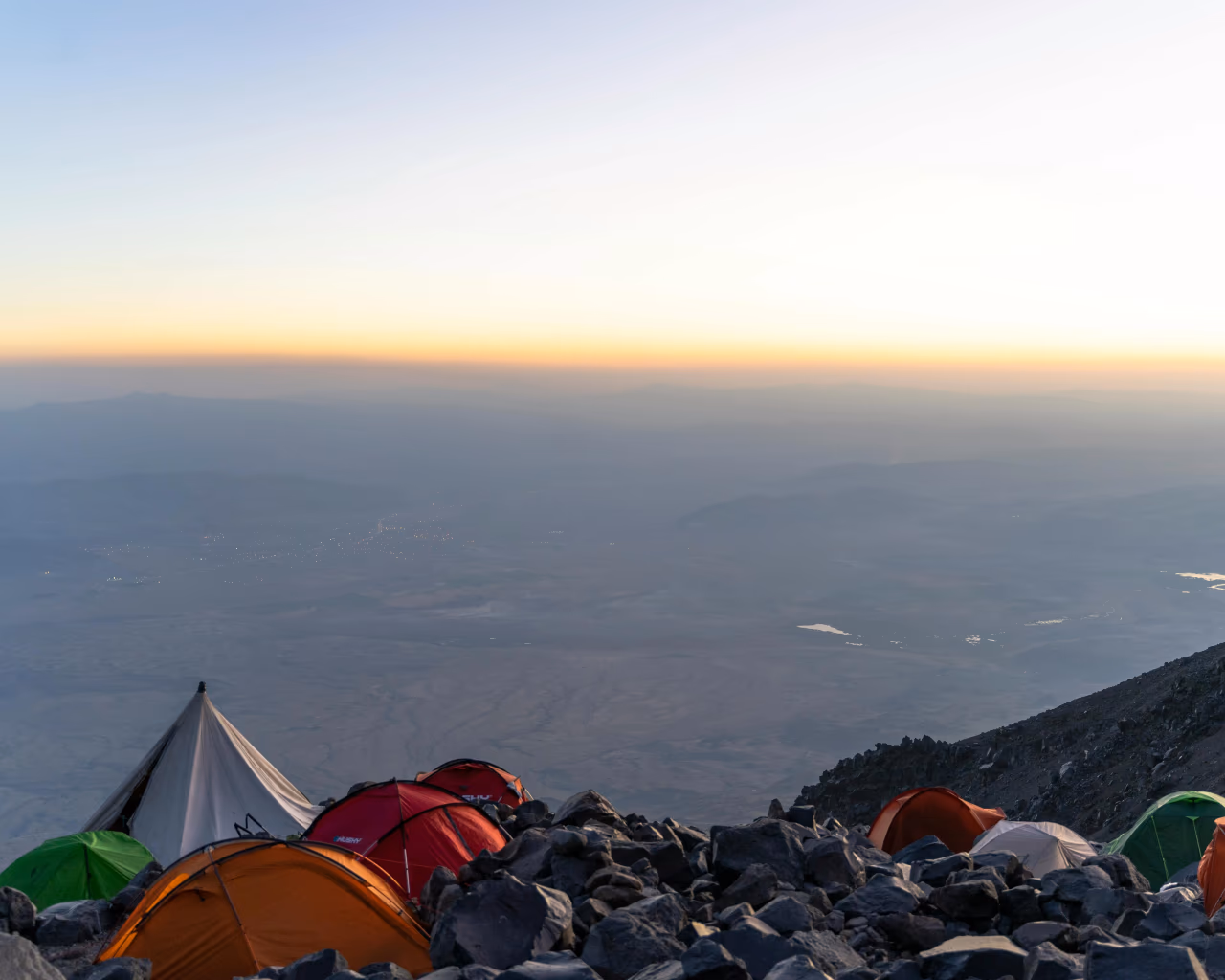 A group of tents pitched on a rocky slope, overlooking a vast landscape at dusk.