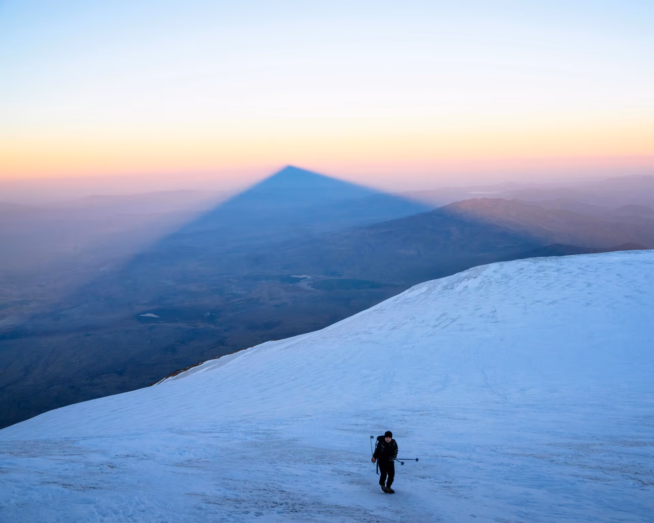 A climber walks on Mount Ararat at sunrise, with the mountain’s triangular shadow in the distance.