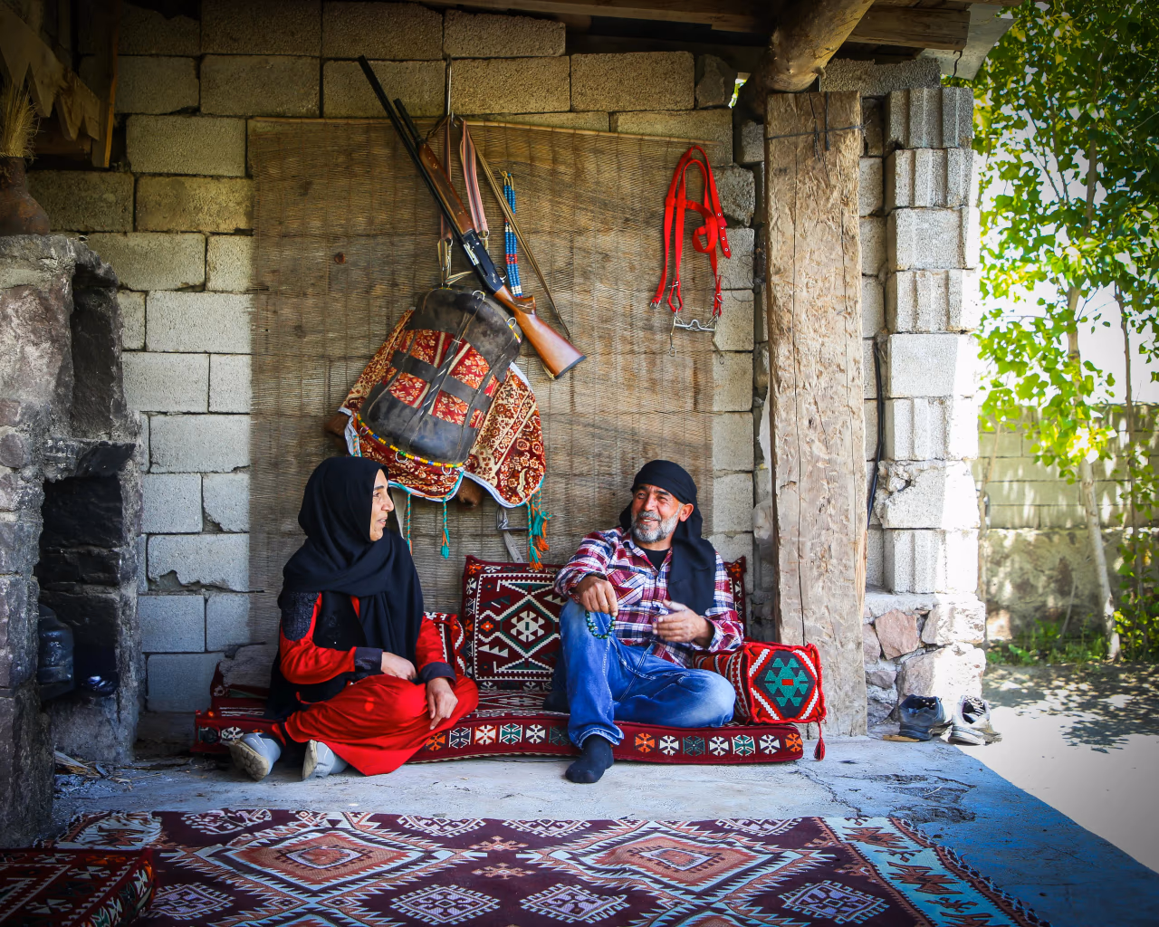 A Kurdish couple sitting on traditional carpets in a rustic home, engaged in conversation.