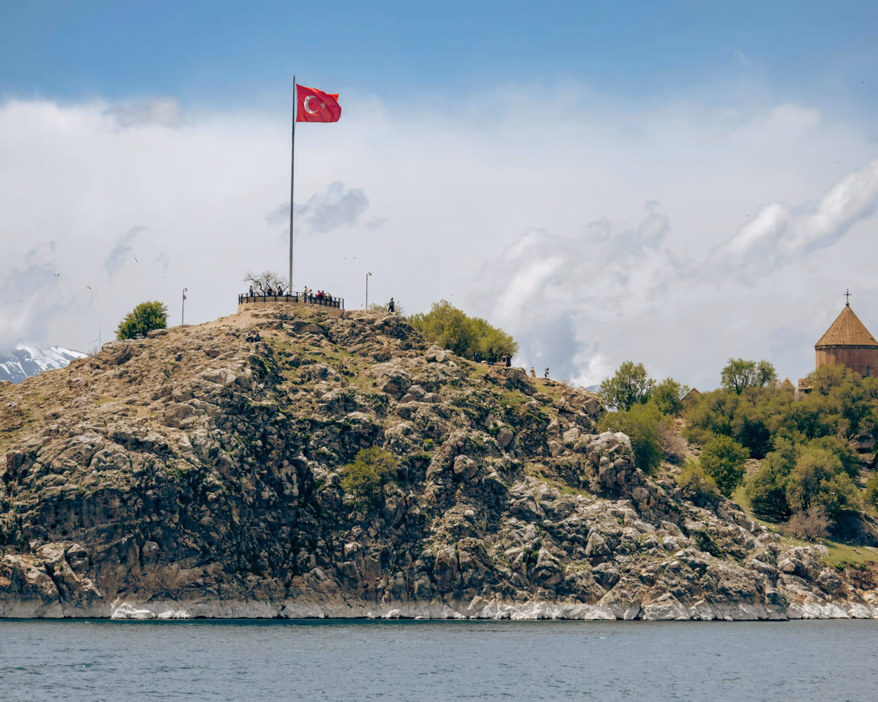A rocky hill with a Turkish flag and a historic church, overlooking a blue lake.