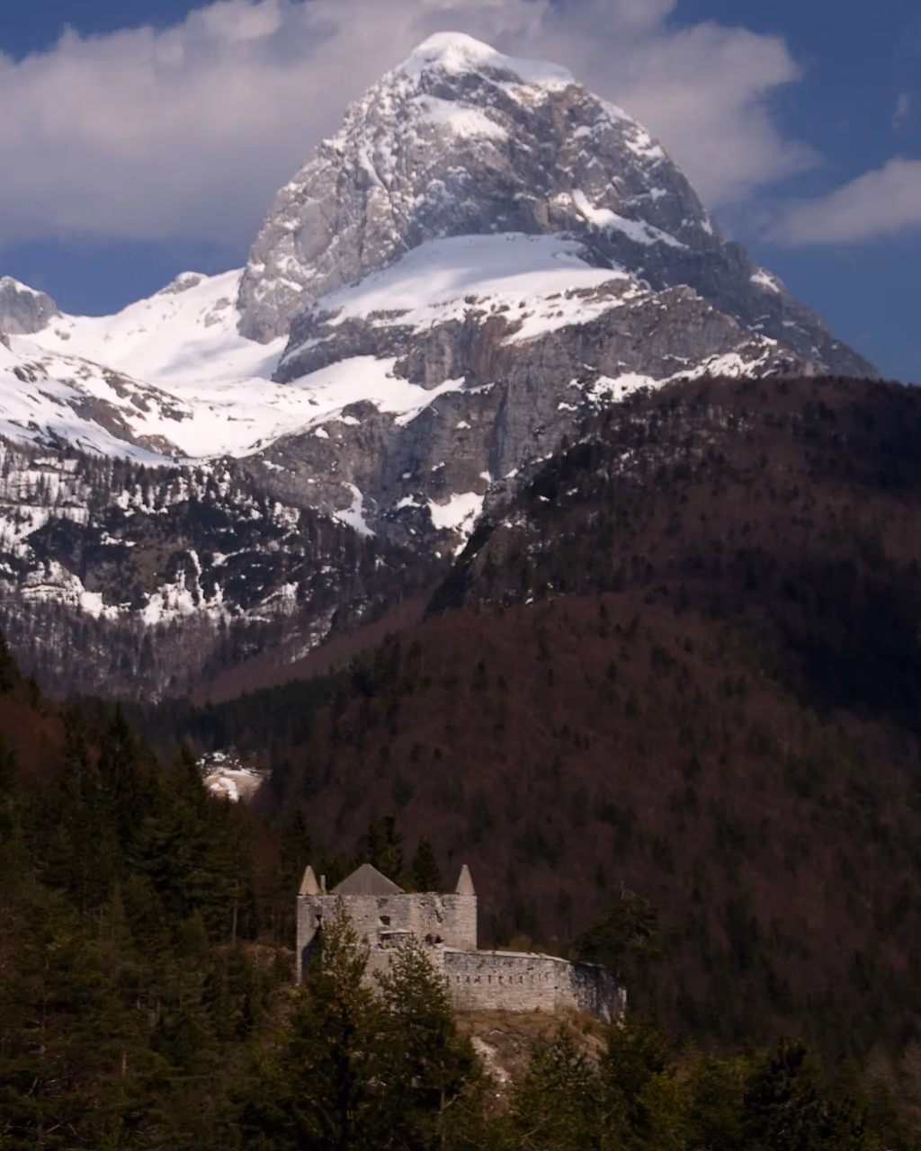 Snow-capped mountain towering above forested hills, with a historic stone building nestled at the base.