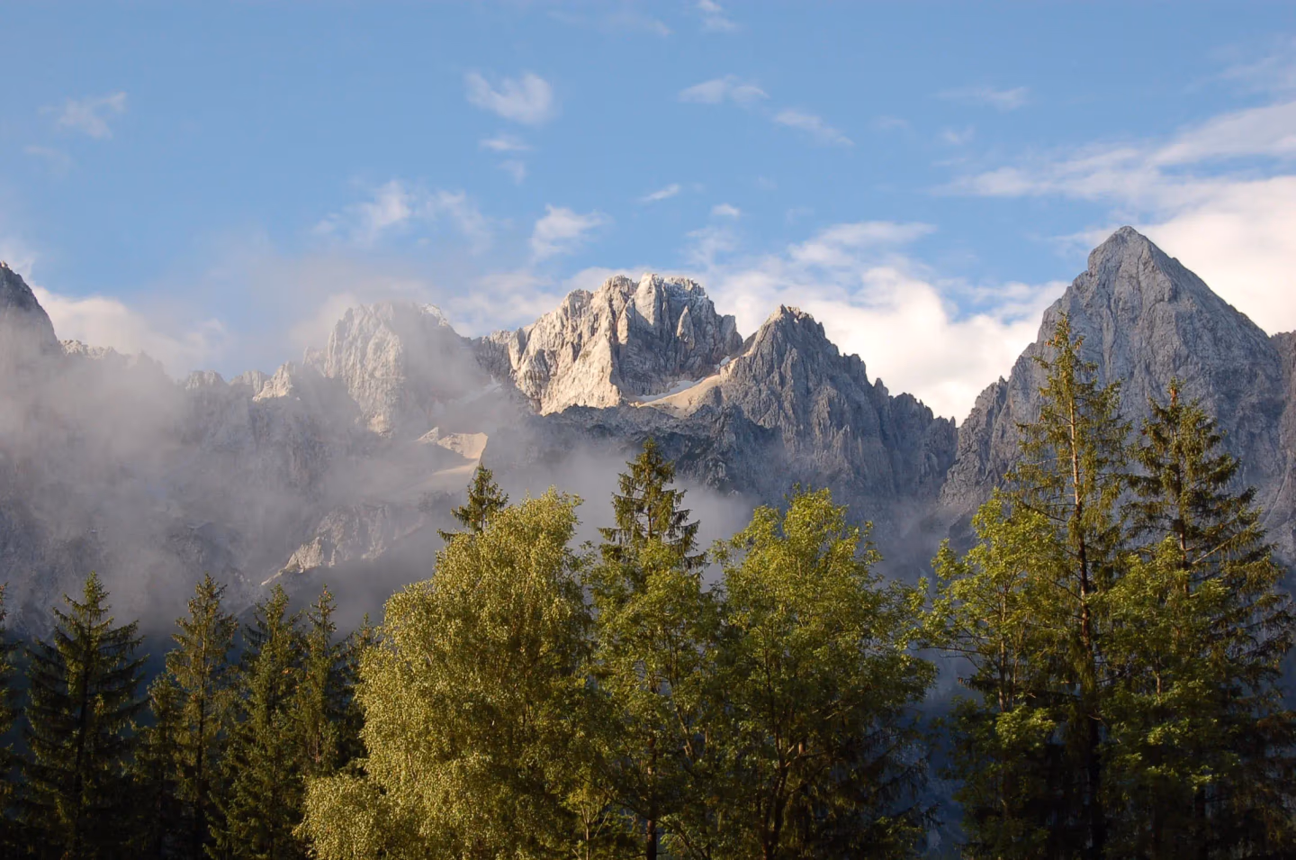 Jagged mountain peaks partially covered in mist, rising above lush green forests and clear blue skies.