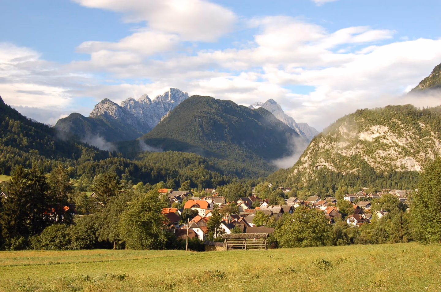 Alpine village with red-roofed houses nestled among green hills and dramatic mountain peaks in the background.