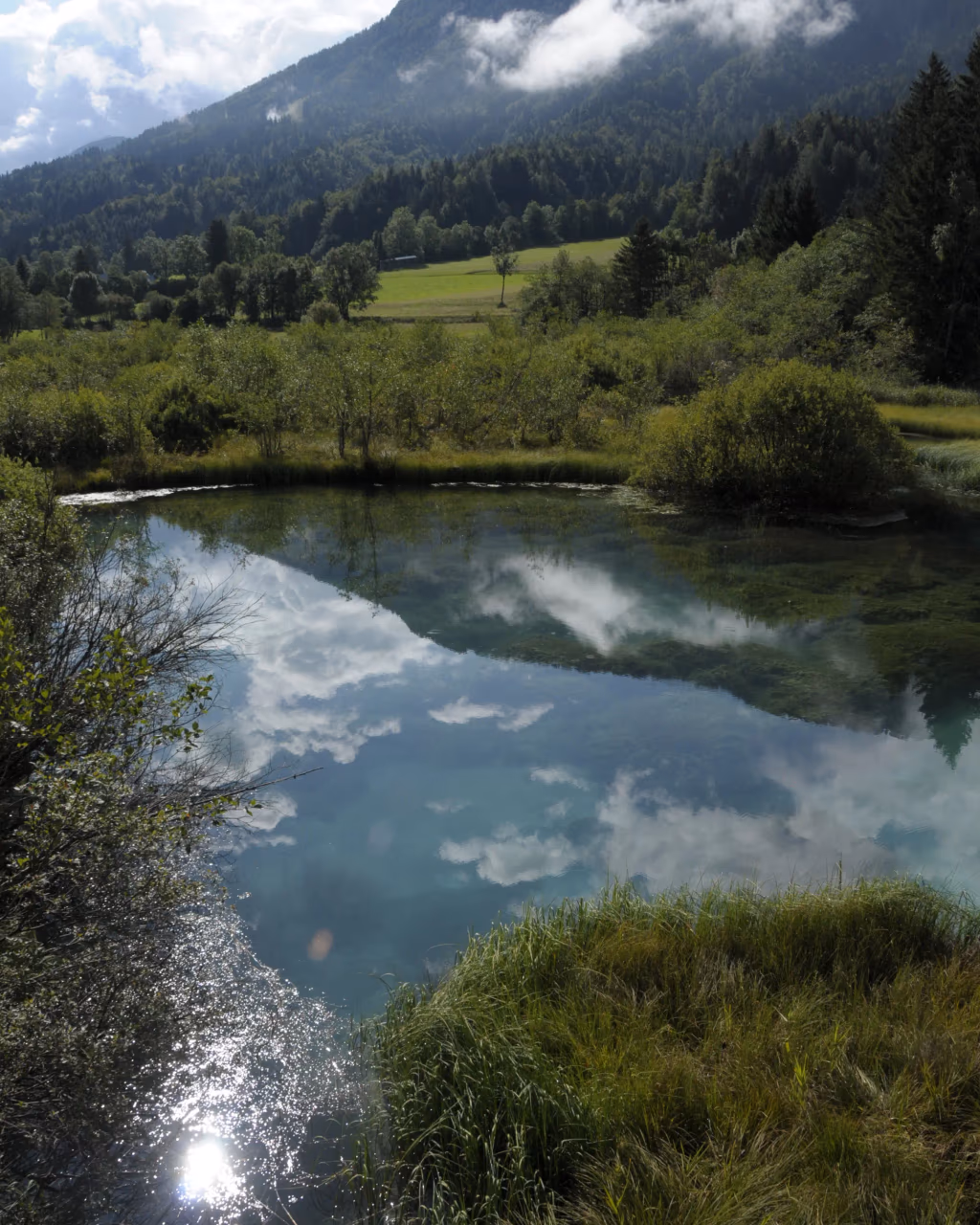 Crystal-clear pond reflecting sky and clouds, surrounded by green meadows and forested hills.