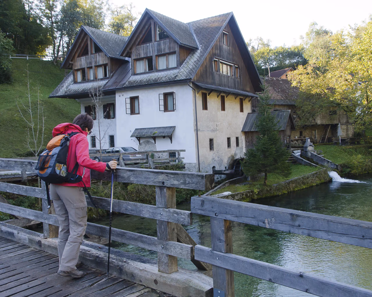 Hiker with trekking poles standing on wooden bridge, looking toward a rustic alpine house and watermill.