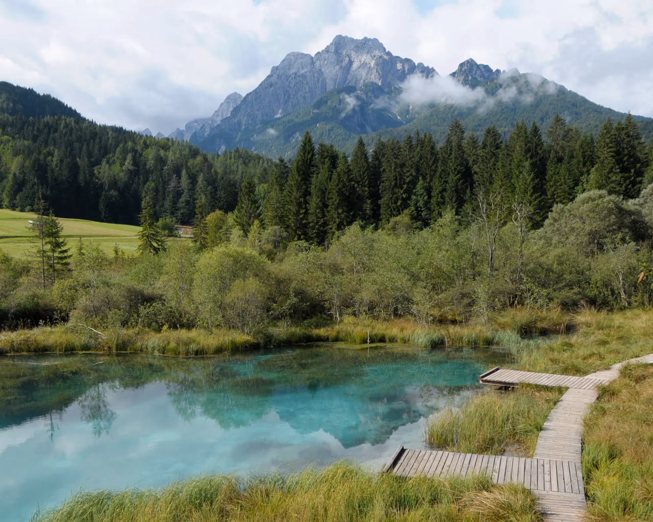Wooden boardwalk leading to a turquoise spring, with mountains and forest forming the scenic backdrop.
