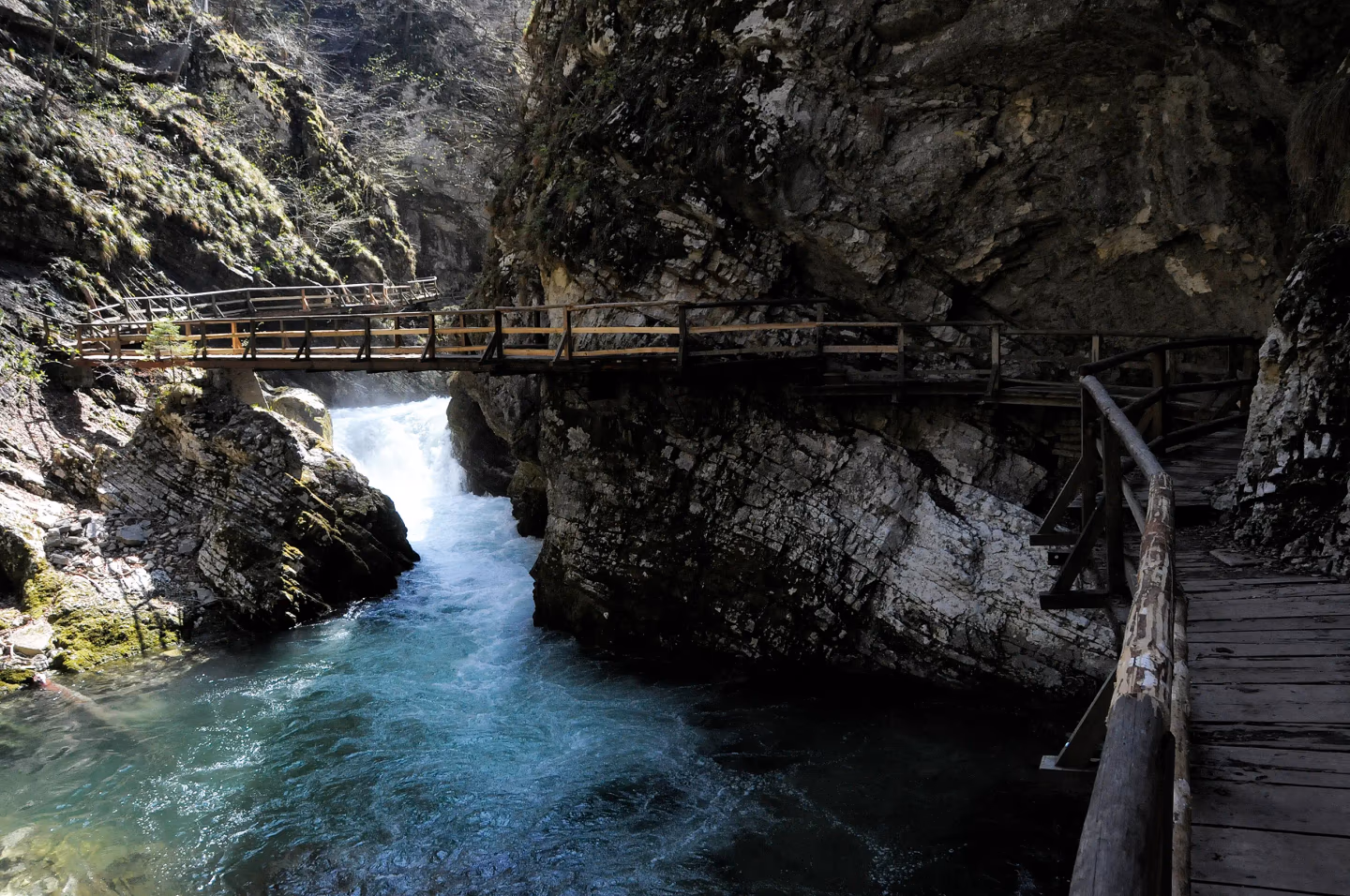 Wooden bridge crossing a turquoise river gorge, with steep rocky cliffs and moss-covered rocks.