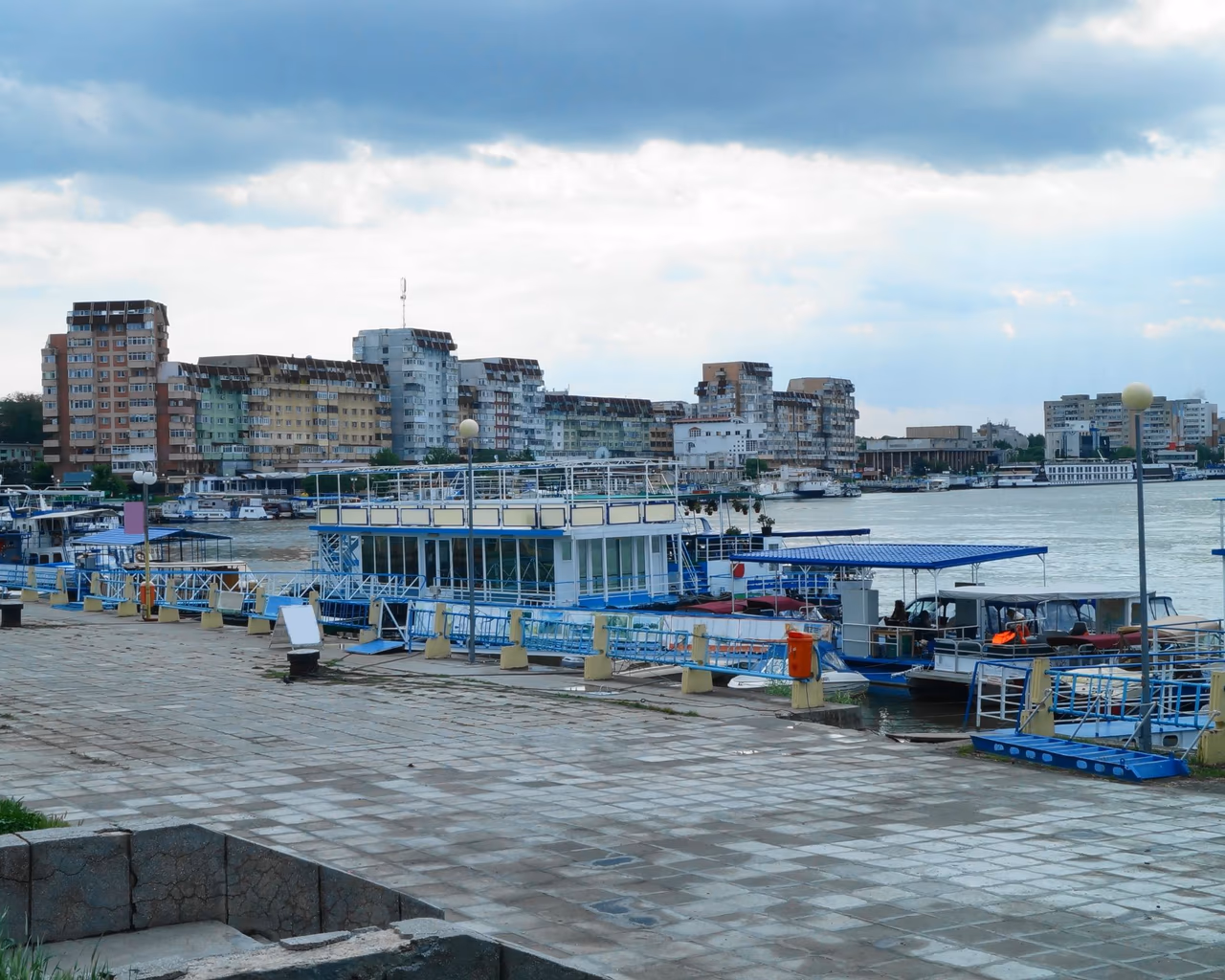Boats docked at a bustling port in Tulcea, Romania, with modern buildings under a cloudy sky in the background.