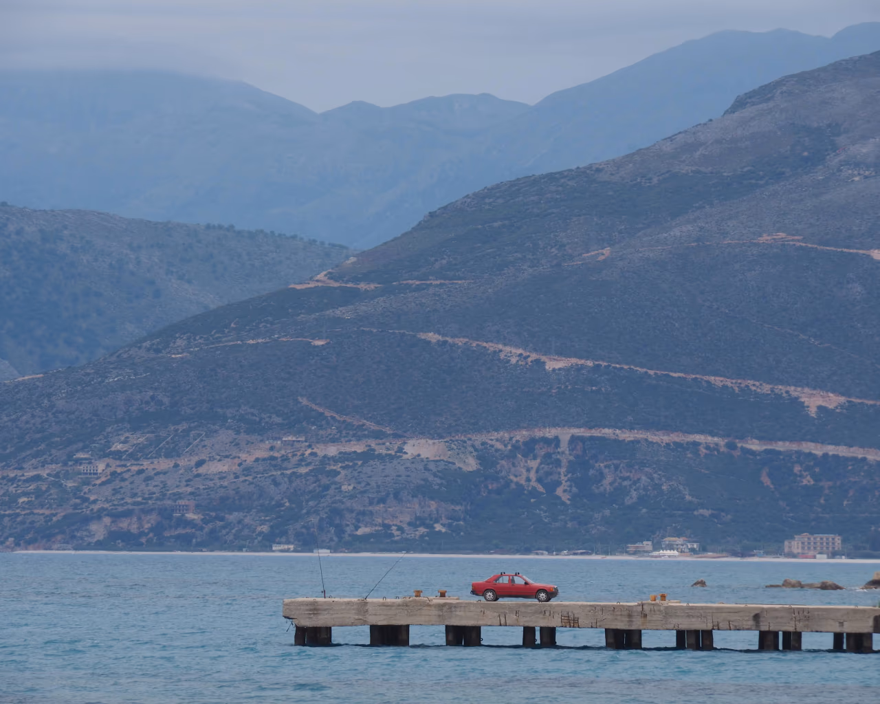 A red car parked at the edge of a pier with rugged mountains in the background along the Albanian Riviera.