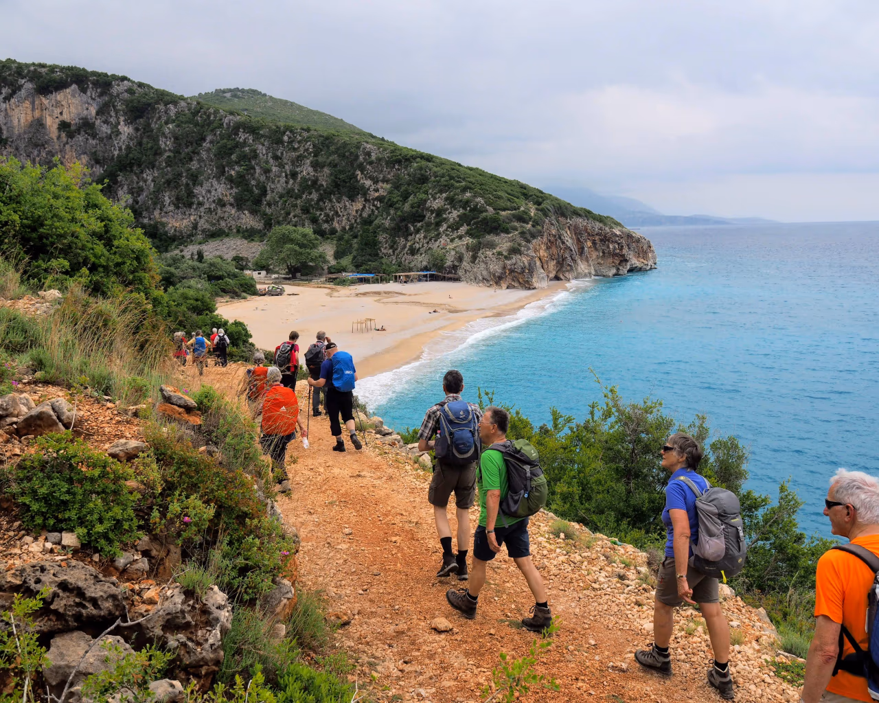 Hikers trekking down a rugged trail towards a secluded sandy beach with turquoise waters along the Albanian coast.