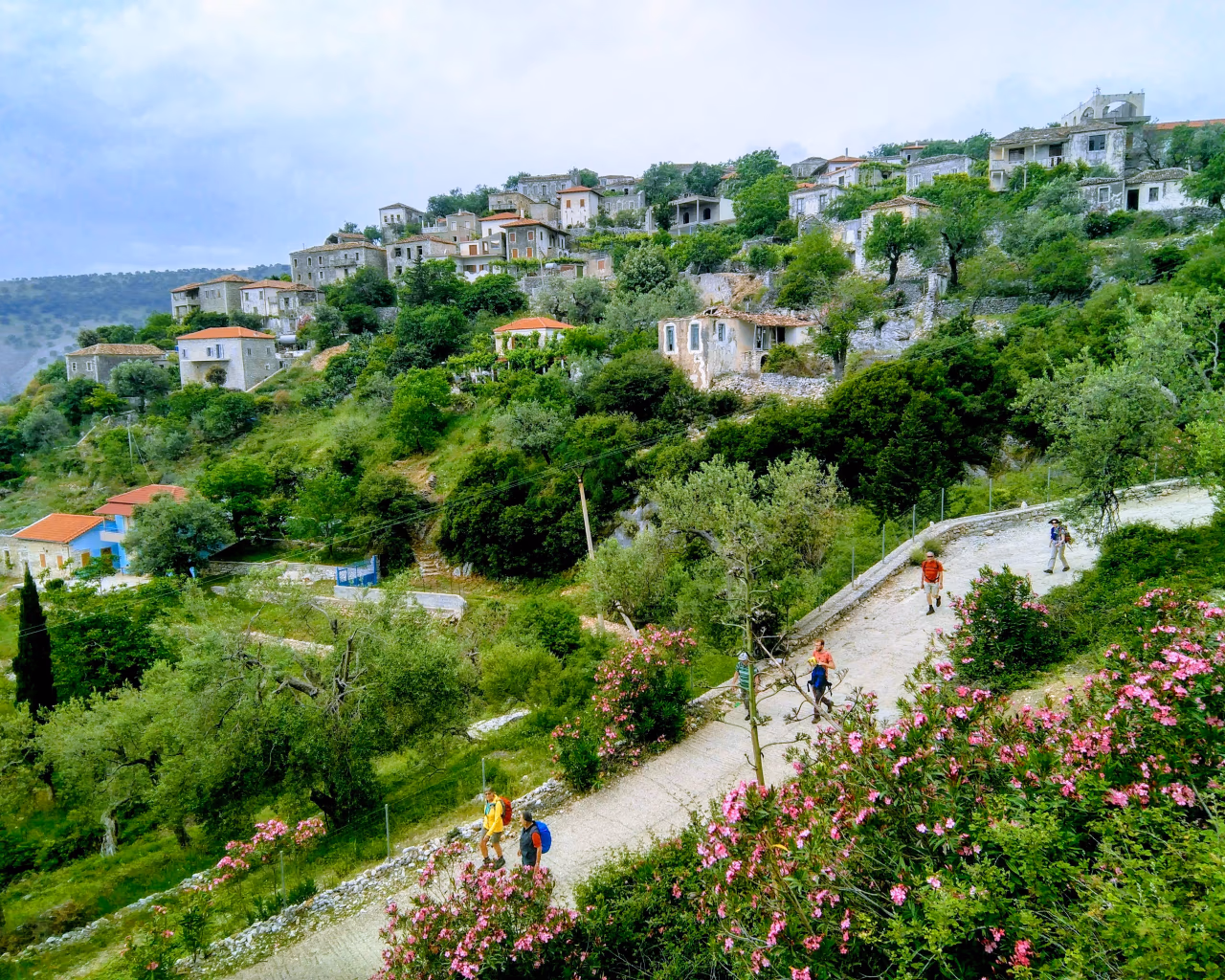 Hikers walking through a lush hillside village with stone houses and colorful flowers in the Albanian countryside.