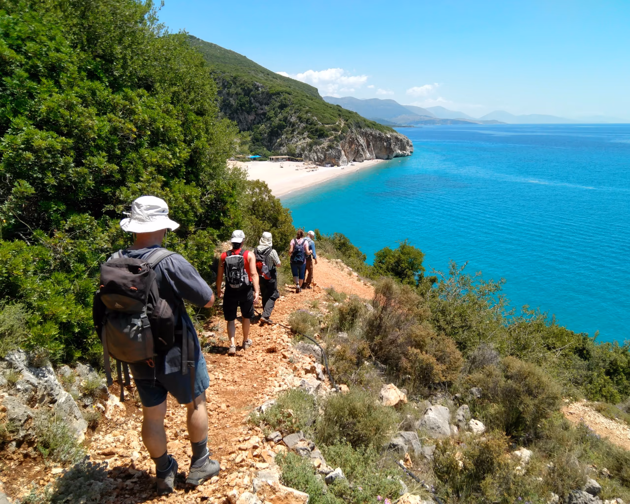 Hikers descend a rugged coastal trail toward a secluded sandy beach with crystal-clear turquoise waters in Albania.