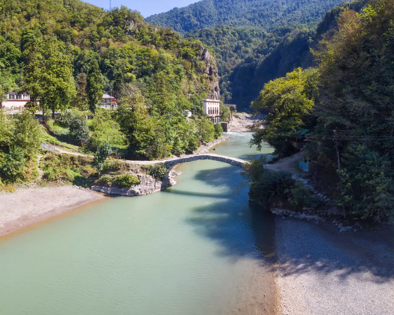 A scenic river winding through lush green mountains in Makhuntseti, Georgia, with a stone bridge crossing over the water.