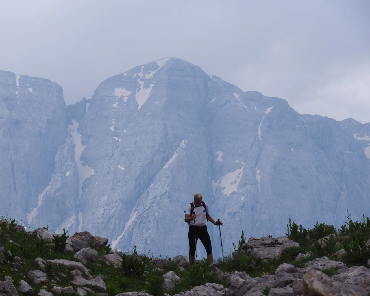 A lone hiker trekking through rocky terrain with towering, snow-dusted peaks in the distance.