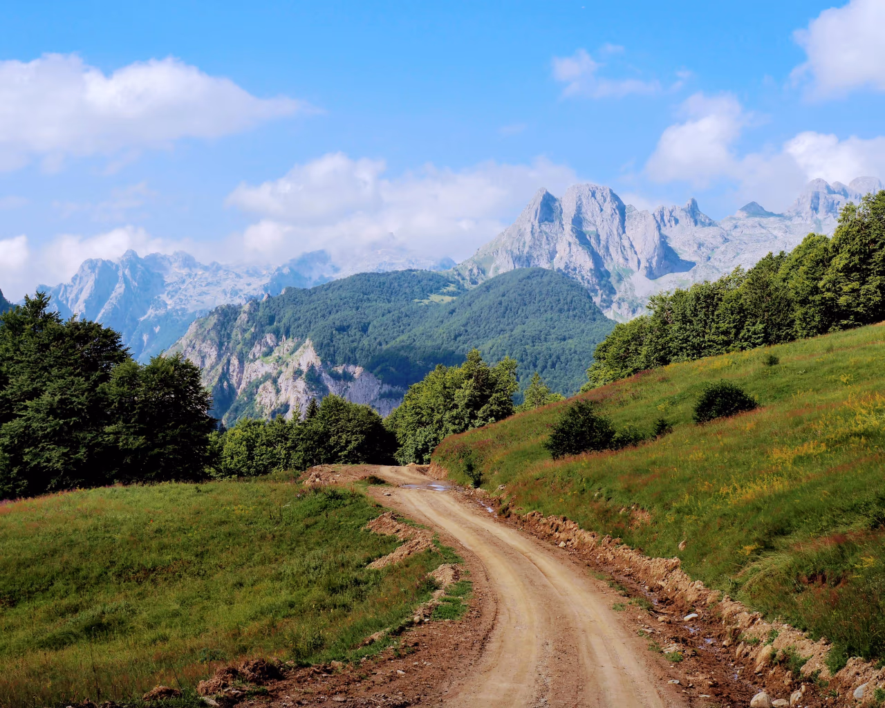 Dirt road winding through green meadows with towering mountains ahead in Albania.