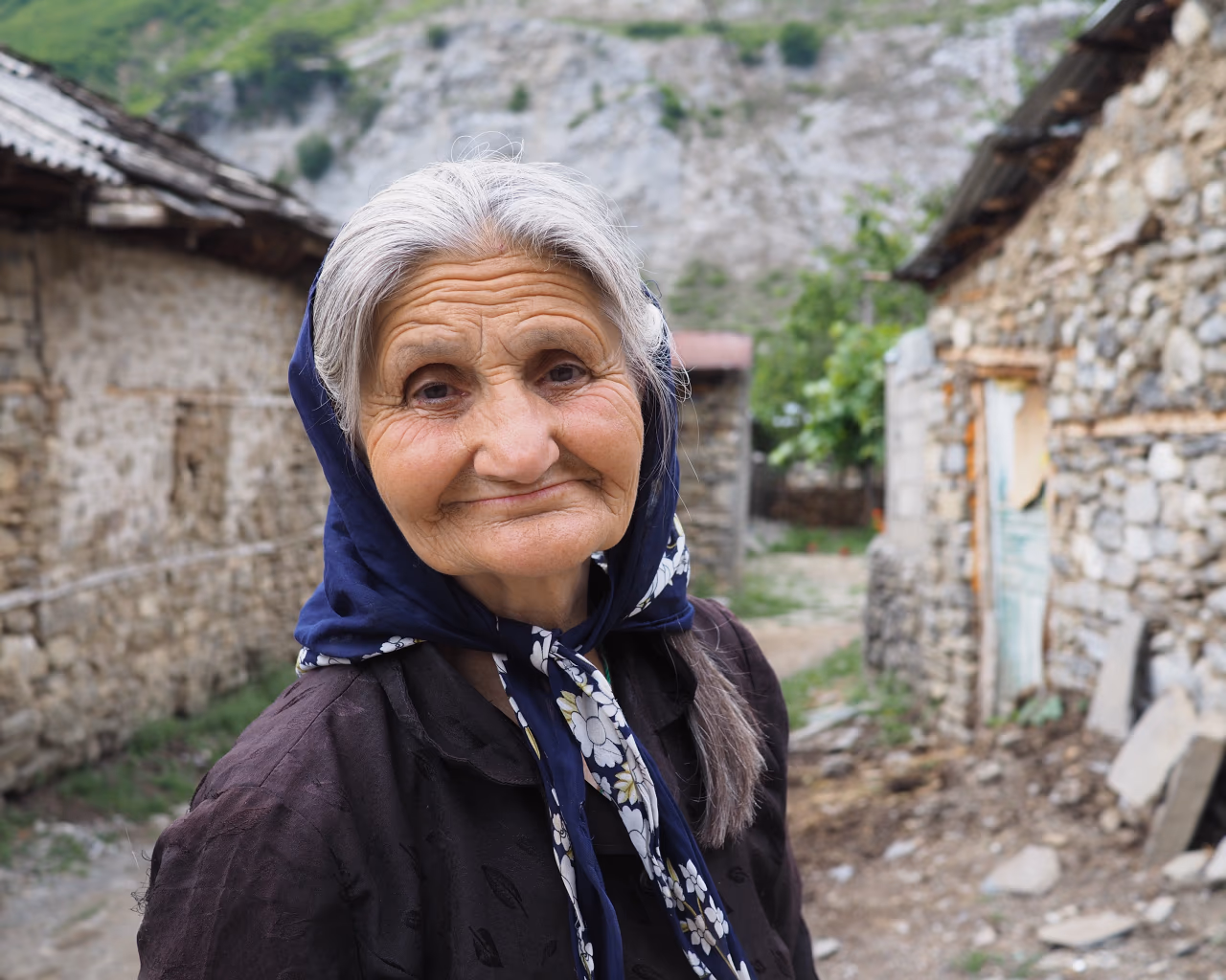 A smiling elderly woman in a blue headscarf stands between rustic stone houses in a mountain village.