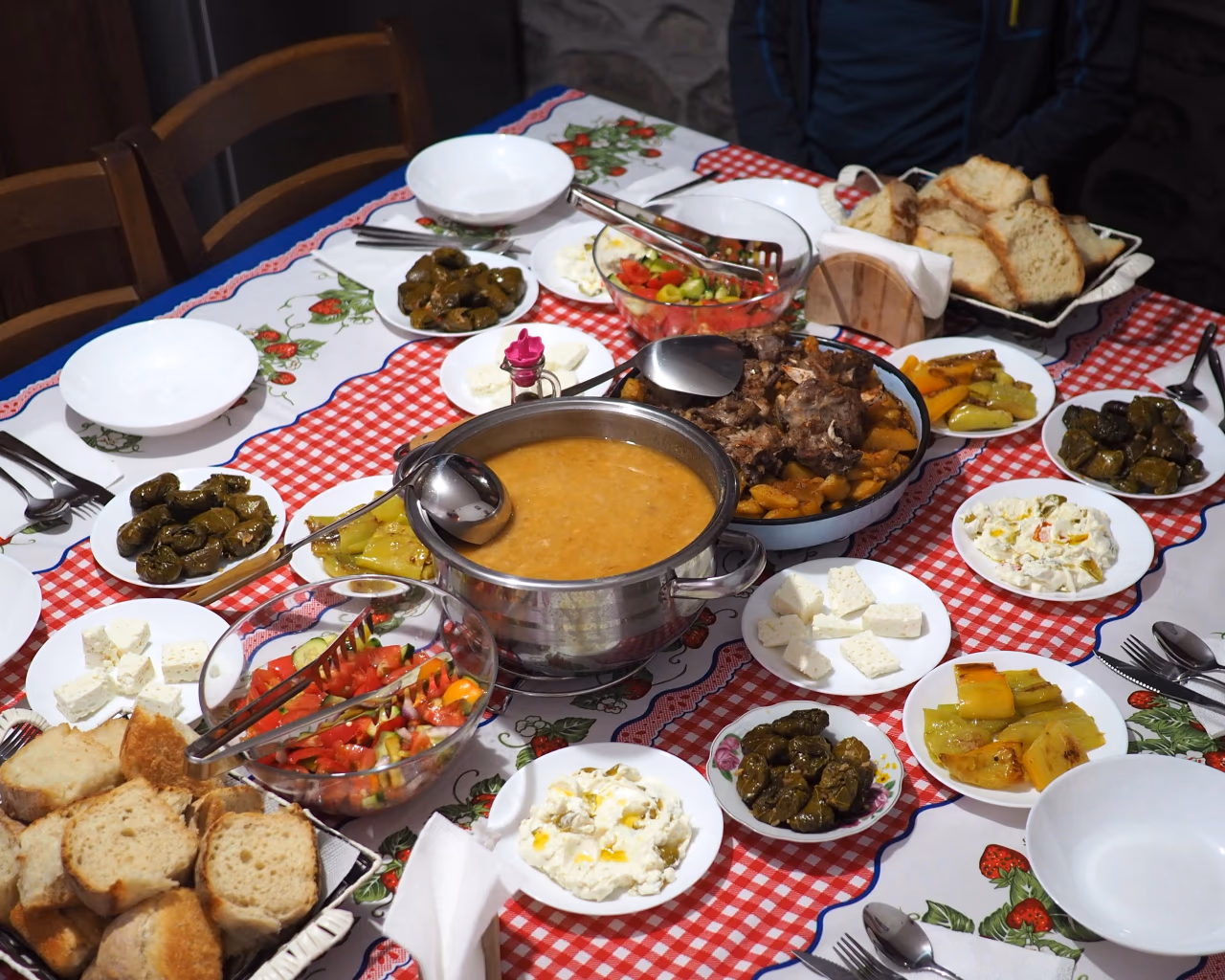 A table with a red-checkered cloth is filled with local dishes, fresh cheese, bread, and stuffed peppers