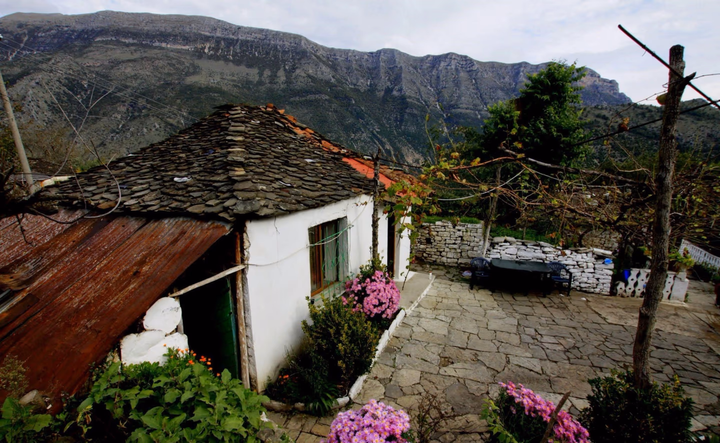 A rustic courtyard with stone pathways, pink flowers, and a small white house against a mountain backdrop.
