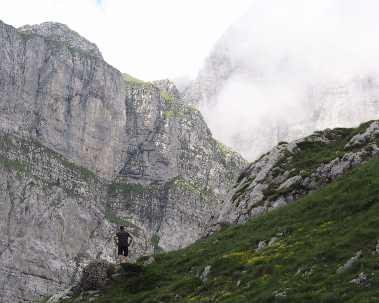 A lone hiker stands atop a grassy ridge, gazing at the towering, mist-covered cliffs of the Albanian Alps.