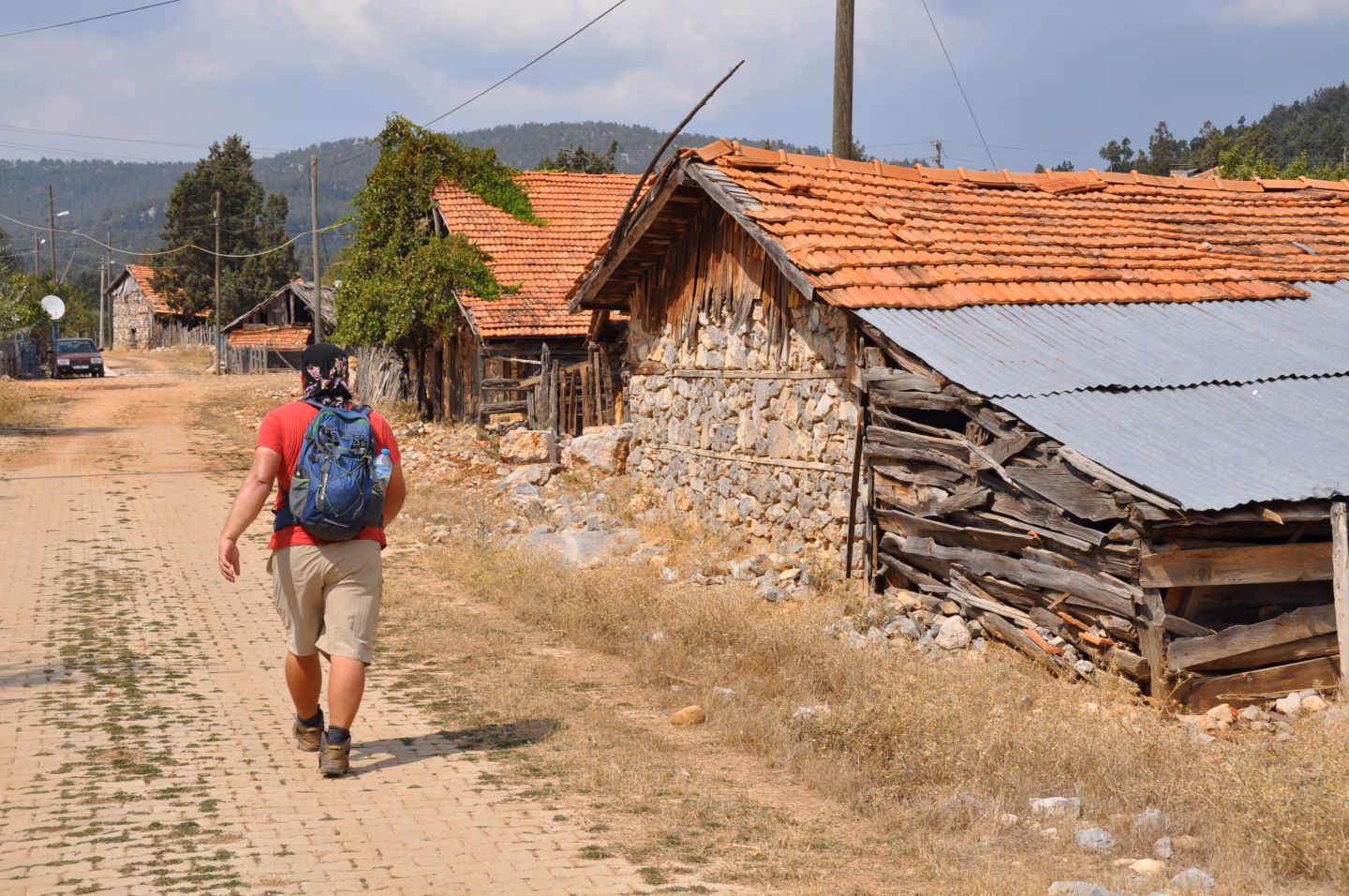 A hiker with a blue backpack walks past old stone houses with red-tiled roofs in a rustic village on the St. Paul Trail.