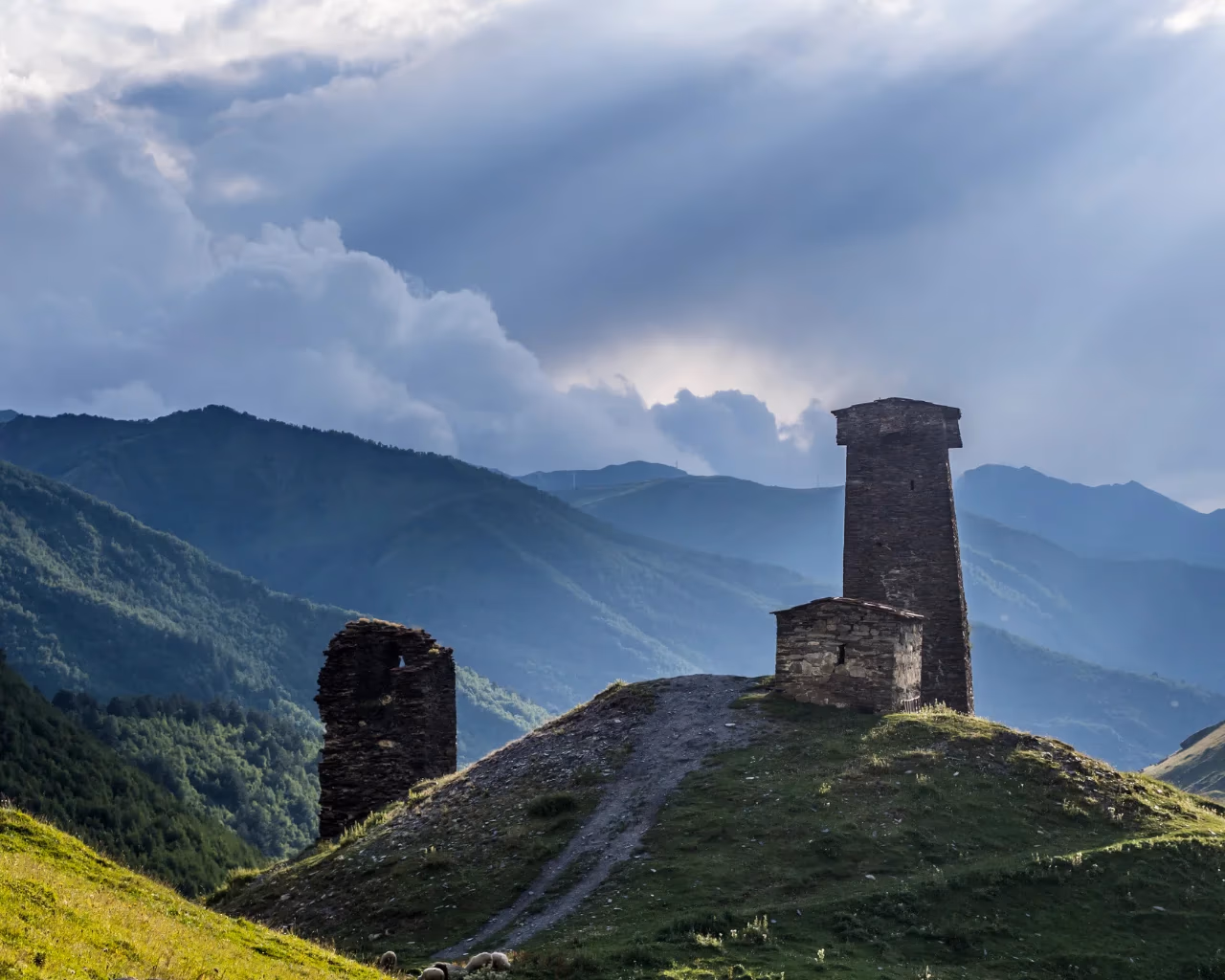 A medieval stone watchtower stands against dramatic clouds, overlooking rolling green hills in Svaneti, Georgia.