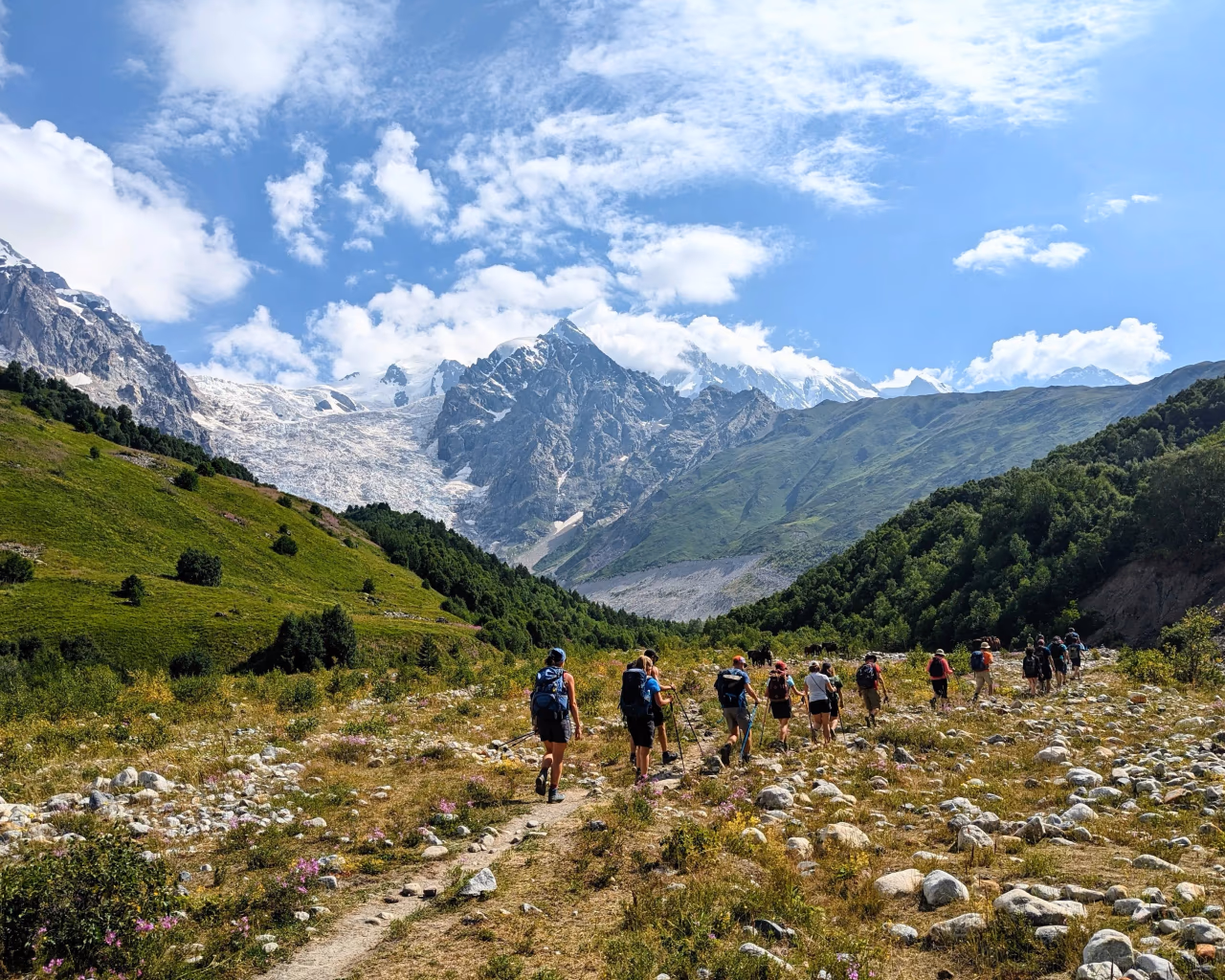 Hikers trekking through lush green meadows in Svaneti, Georgia, with traditional medieval Svan stone towers and snow-capped Caucasus peaks in the background.