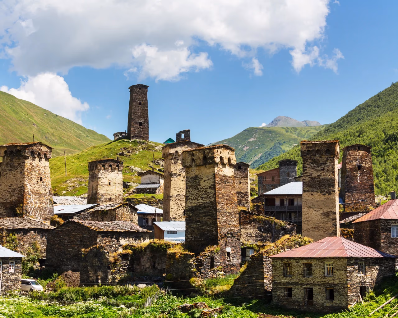 Stone defensive towers dominate the mountain village of Ushguli, Georgia, under a bright blue sky with scattered clouds.