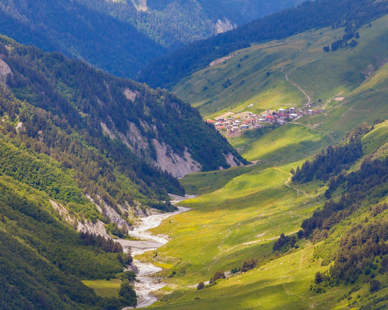 A small Georgian village with red-roofed houses is nestled among lush green hills and a winding river in Svaneti.