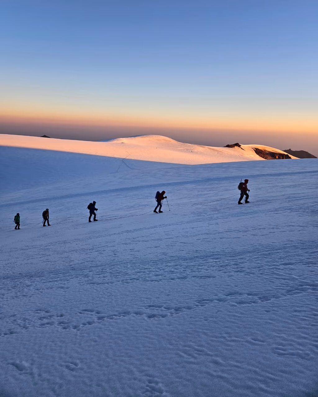 Rope team hikes across Mount Kazbek glacier under soft pastel sunrise sky.
