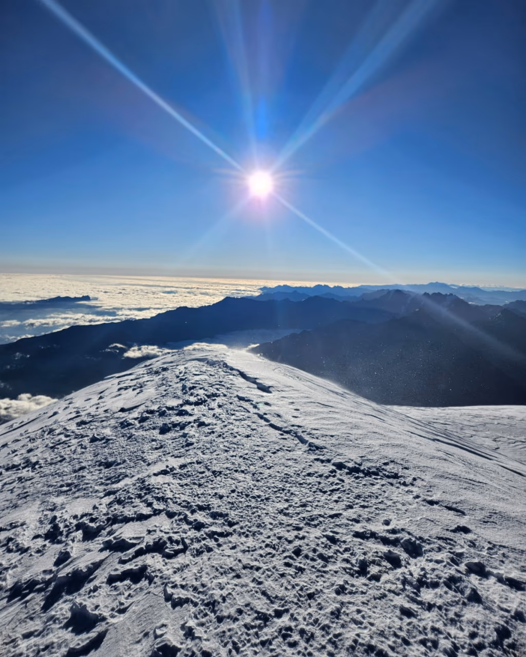 Snow-covered mountain summit rising above a sea of clouds under bright sunburst in clear blue sky