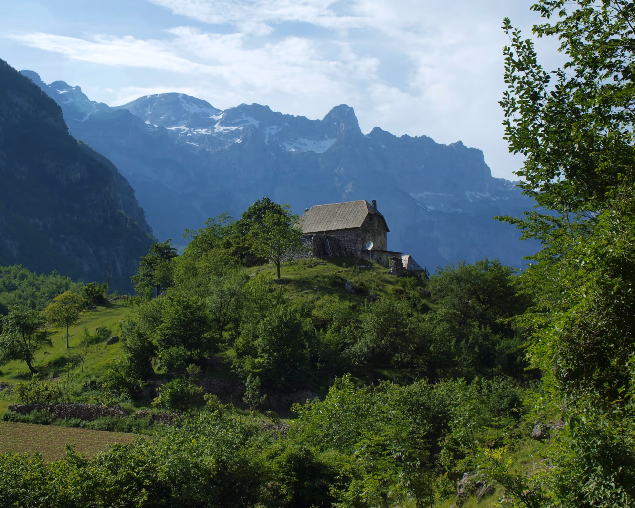 Remote stone house on a lush hill in Theth, Albania, with towering alpine peaks in the background.