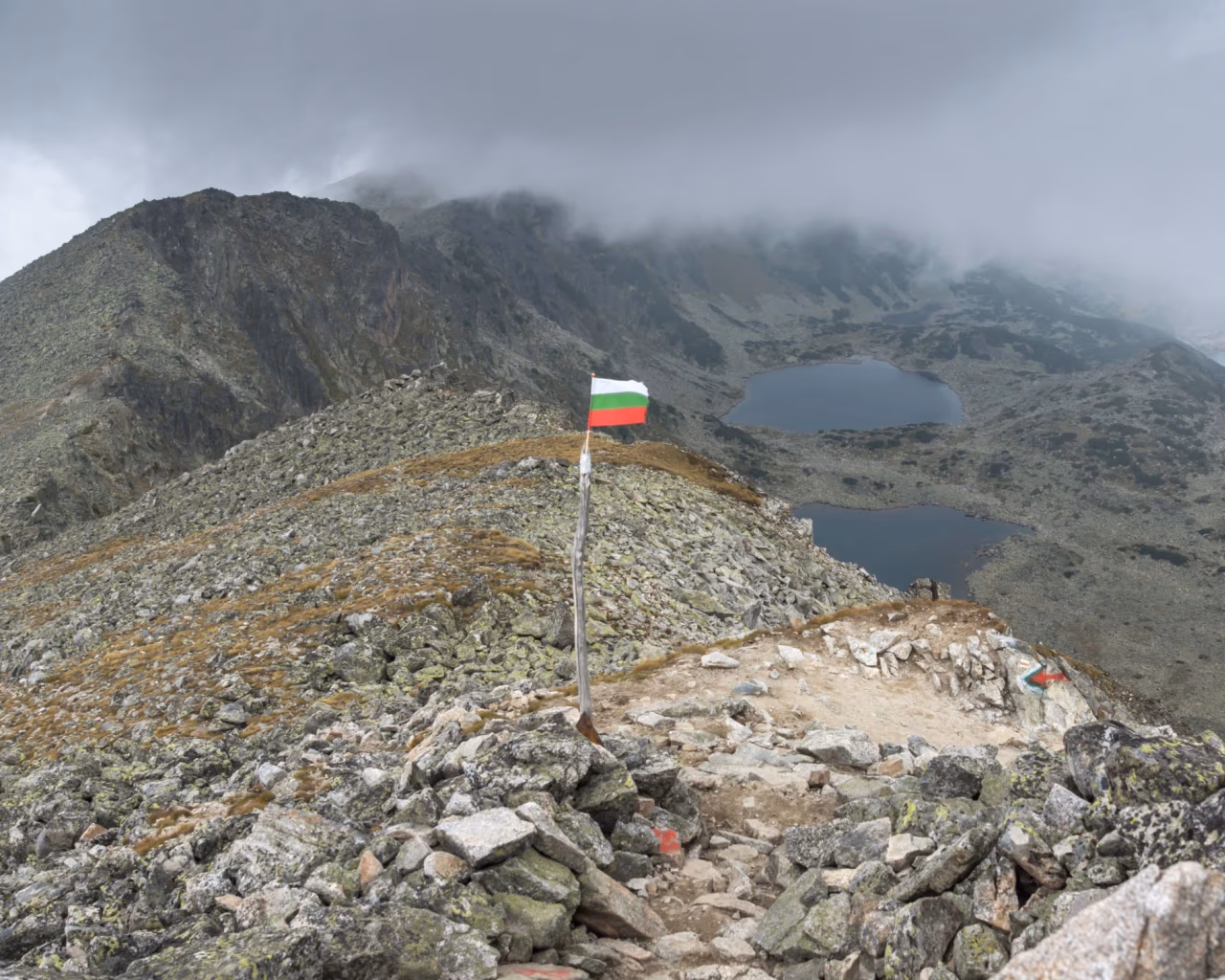 Overcast view from the summit of Musala Peak, Bulgaria, a Bulgarian flag standing against a misty mountain backdrop.