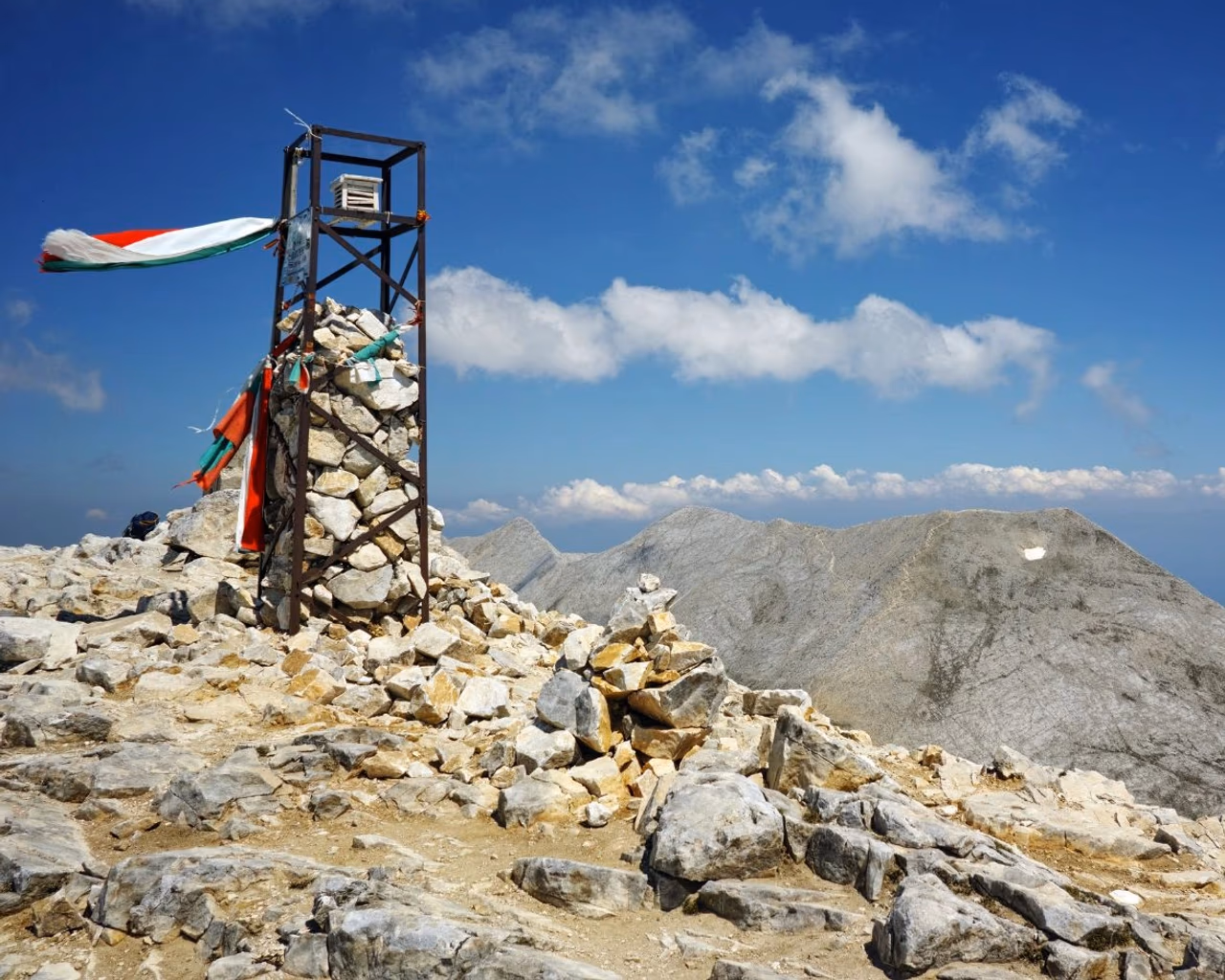 Trig point cairn draped with Bulgarian flag on Mount Vihren summit overlooking rocky Pirin peaks