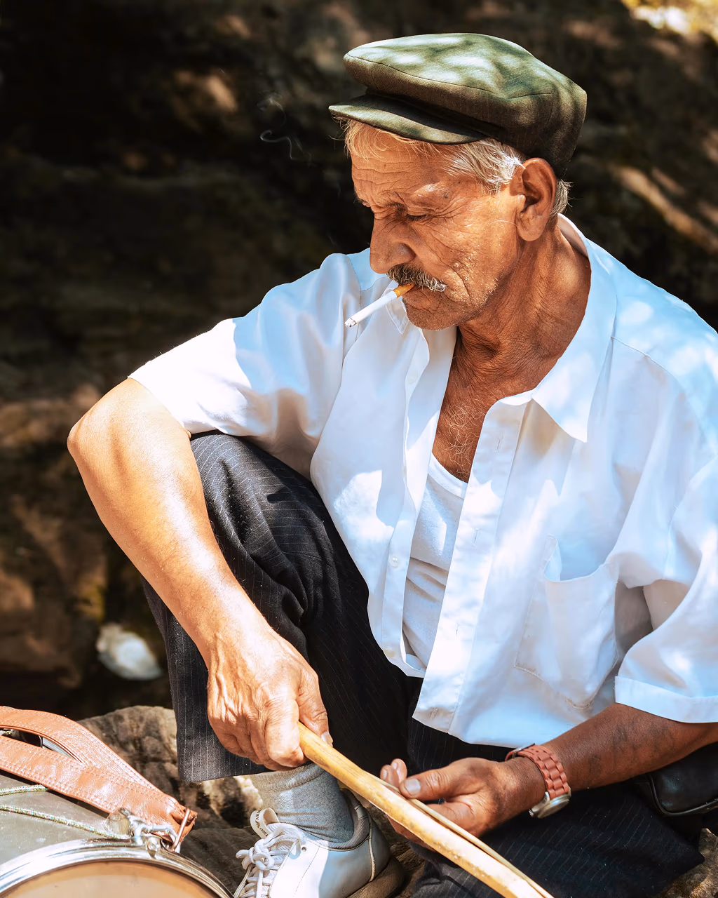 Elderly man in a white shirt and cap playing drums outdoors in Albania, smoking a cigarette.