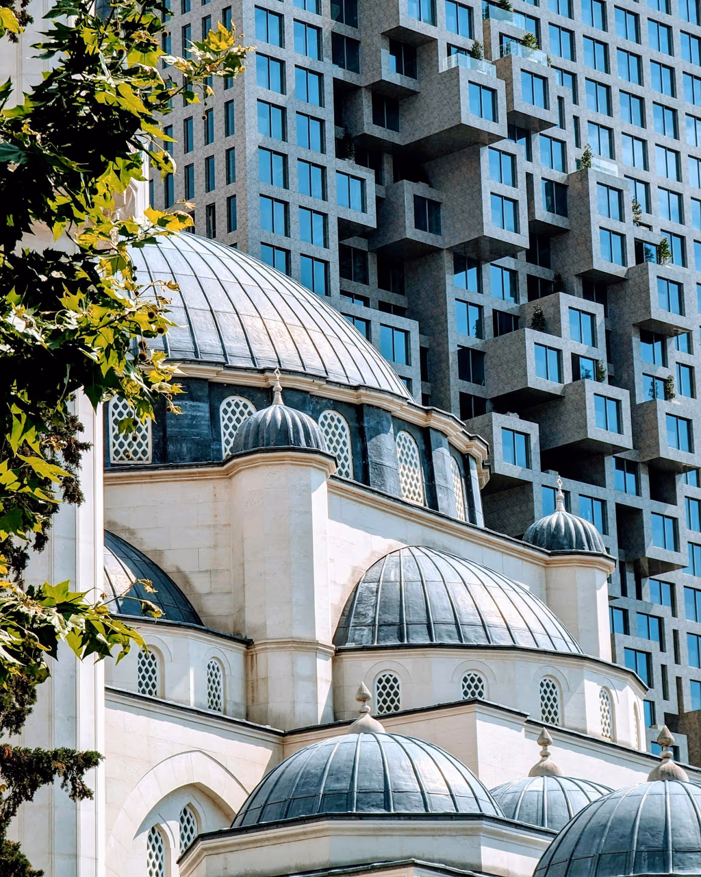 A modern geometric high-rise building behind an Ottoman-style mosque with domed rooftops in Tirana, Albania.