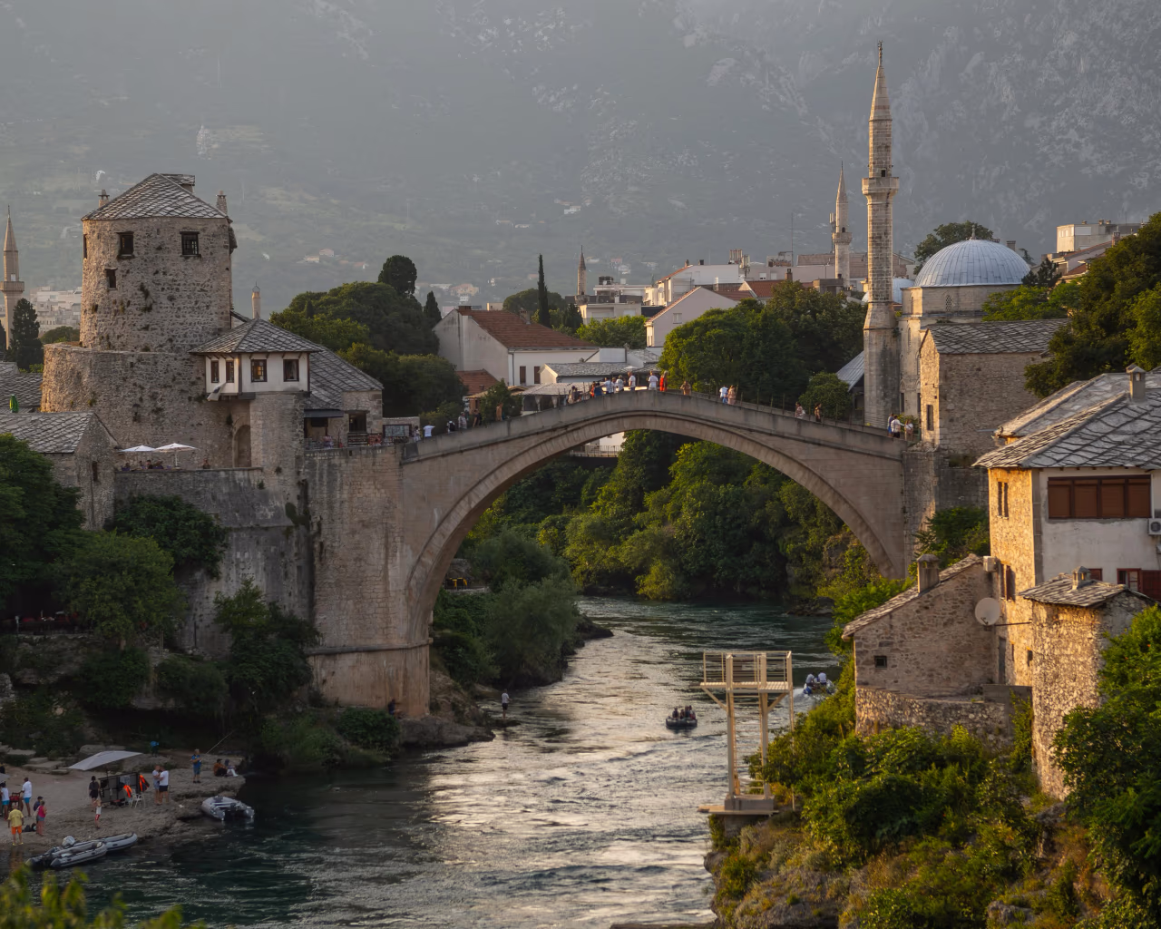 The historic Stari Most bridge in Mostar, Bosnia, at sunset, with people walking across and boats in the river below.