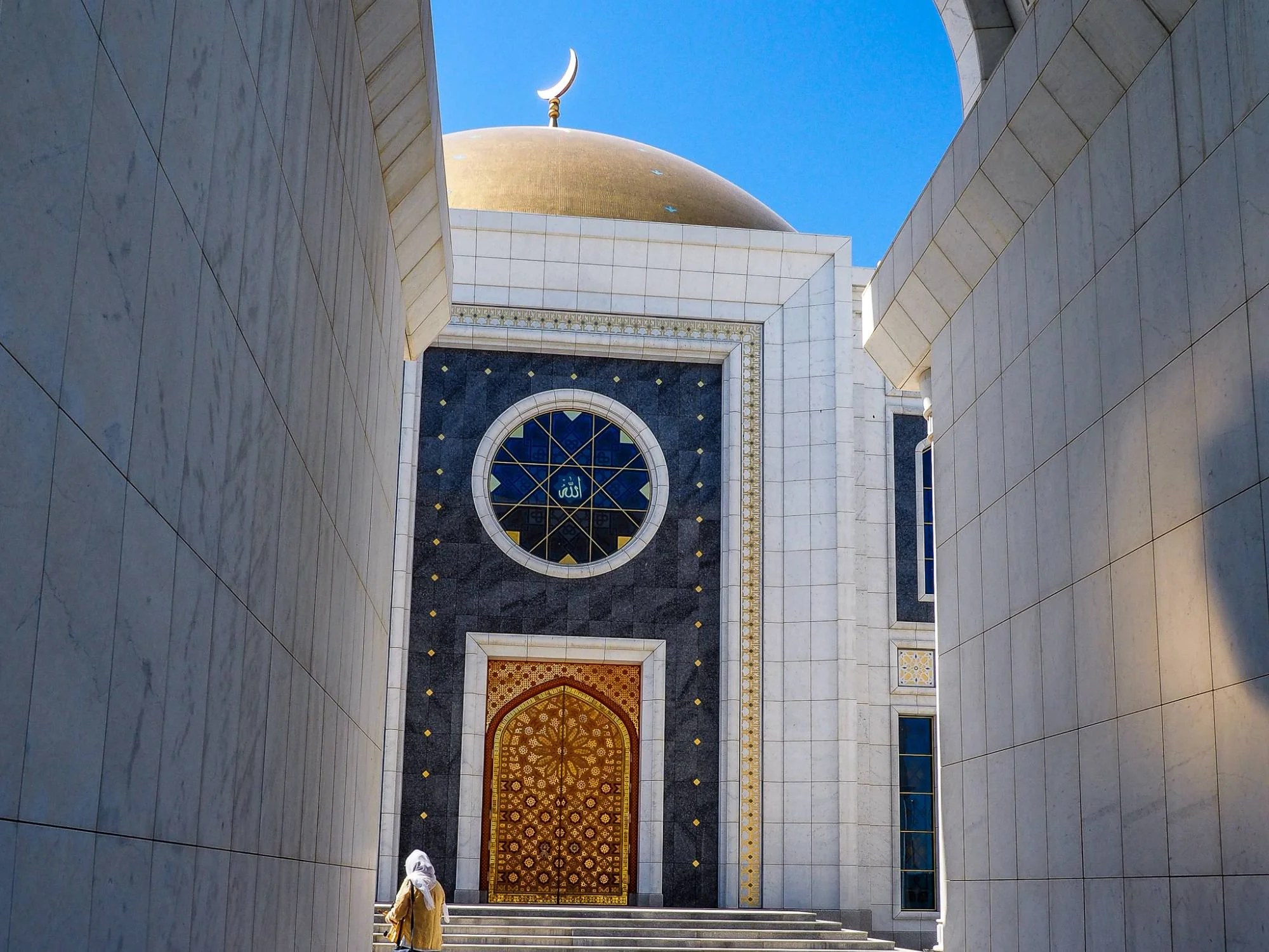 Visitor climbing the steps to a golden-domed mosque with gilded doors and a stained-glass rose window, Turkmenistan