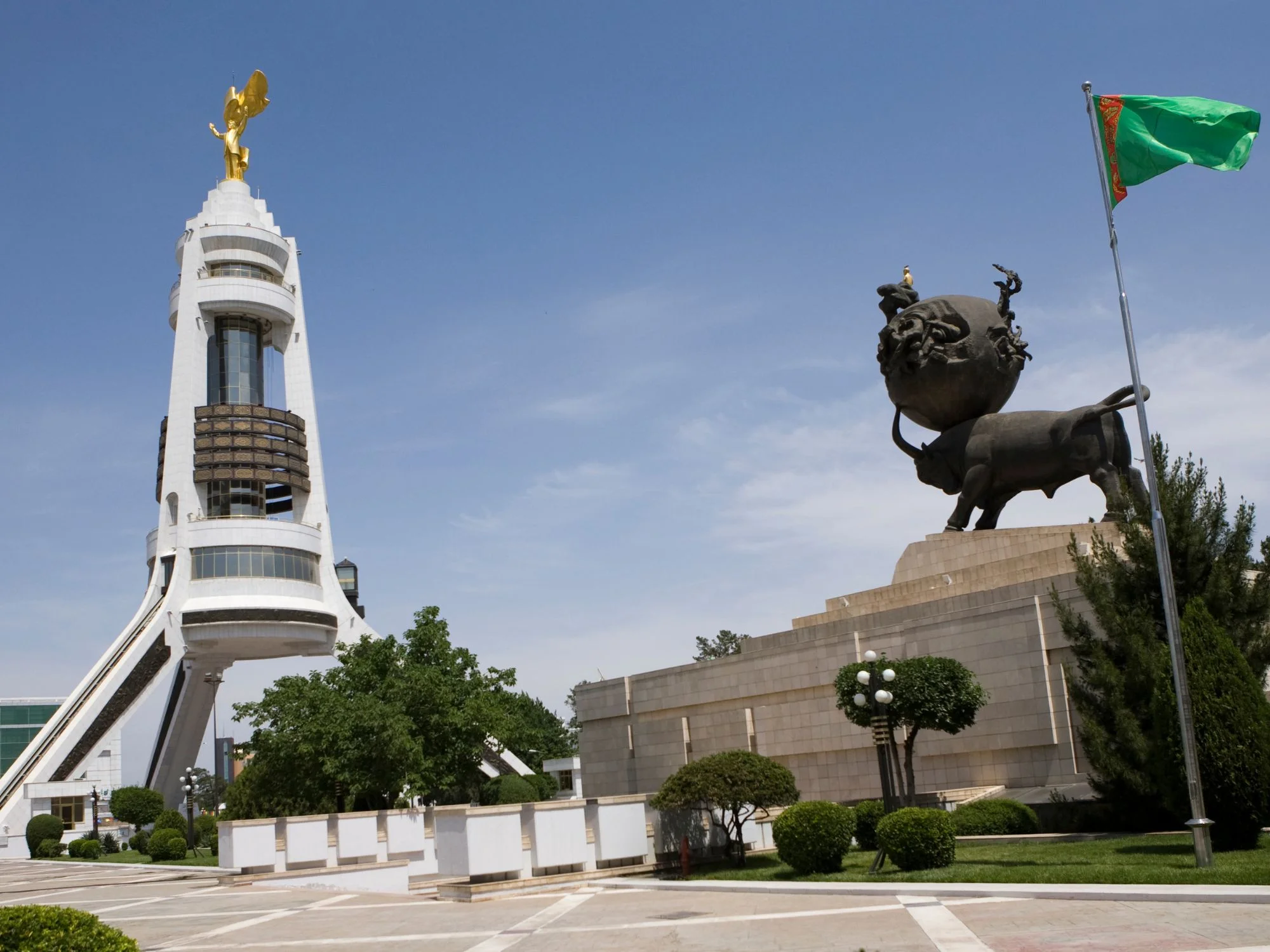 White tripod tower topped by a gilded statue beside a bronze bull-and-globe monument, Ashgabat