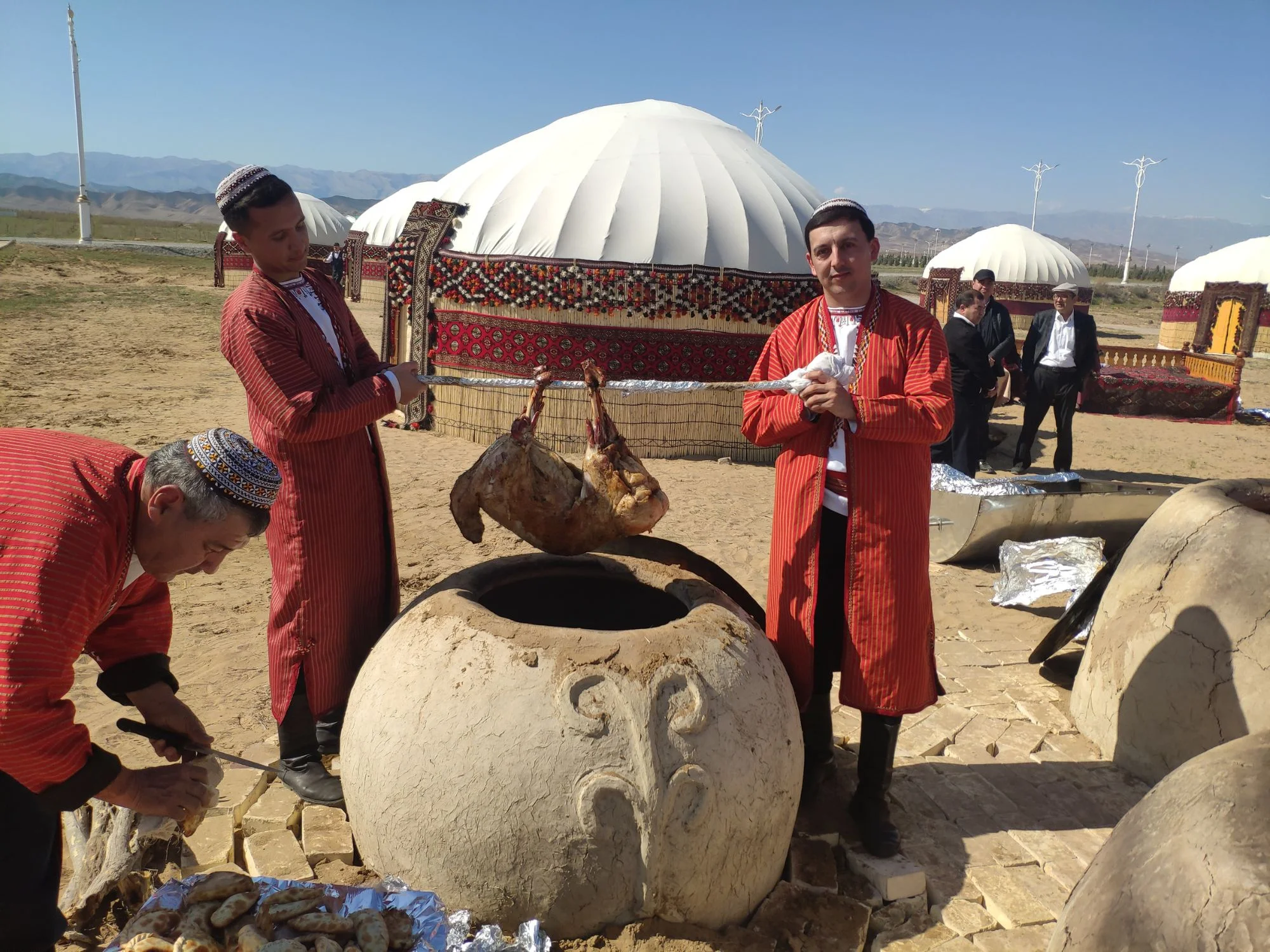 Men in red robes preparing flatbread and roast lamb at a clay tandoor beside festival yurts