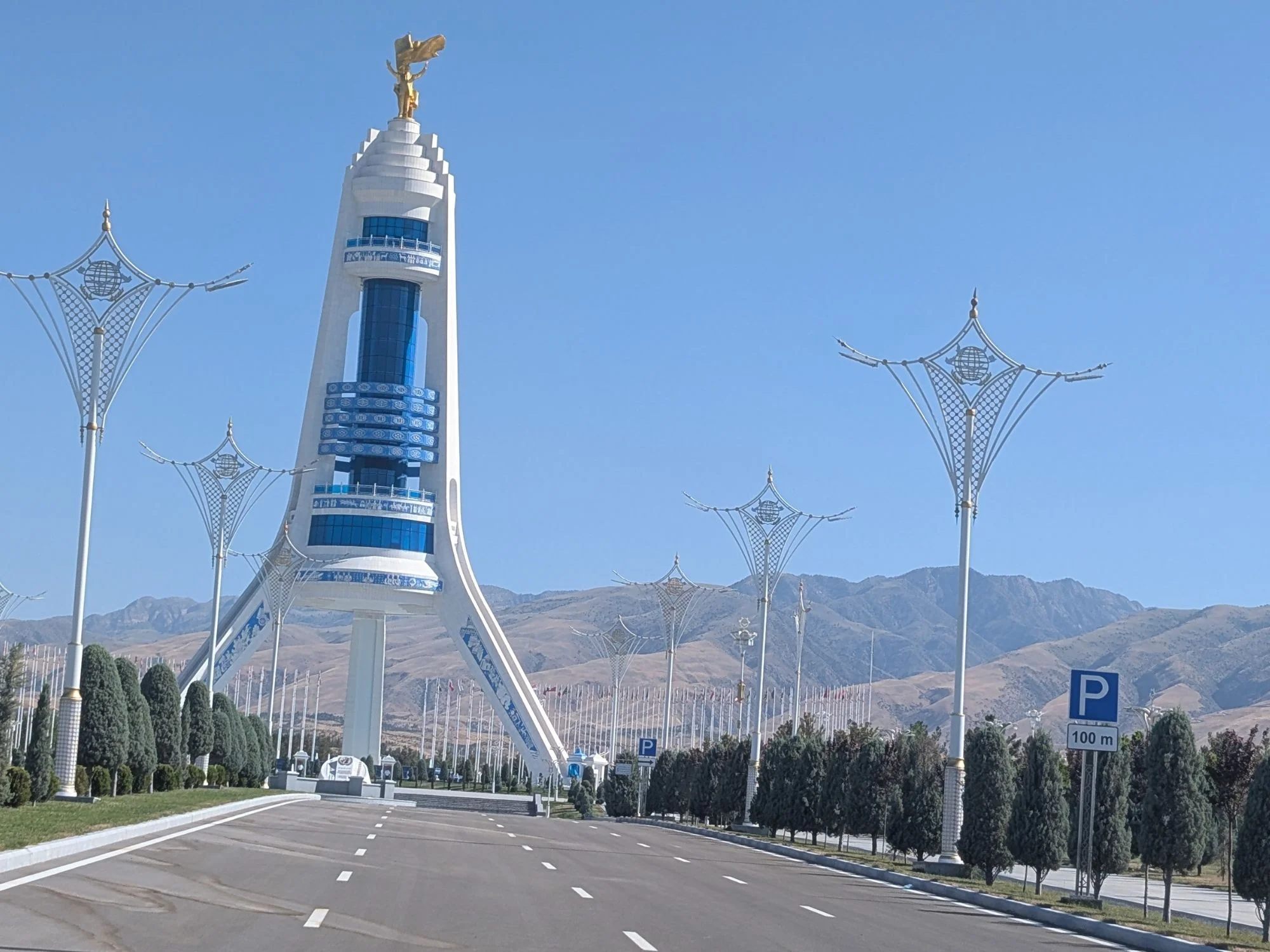 Tall white monument topped by a gilded statue on an empty avenue below arid mountains