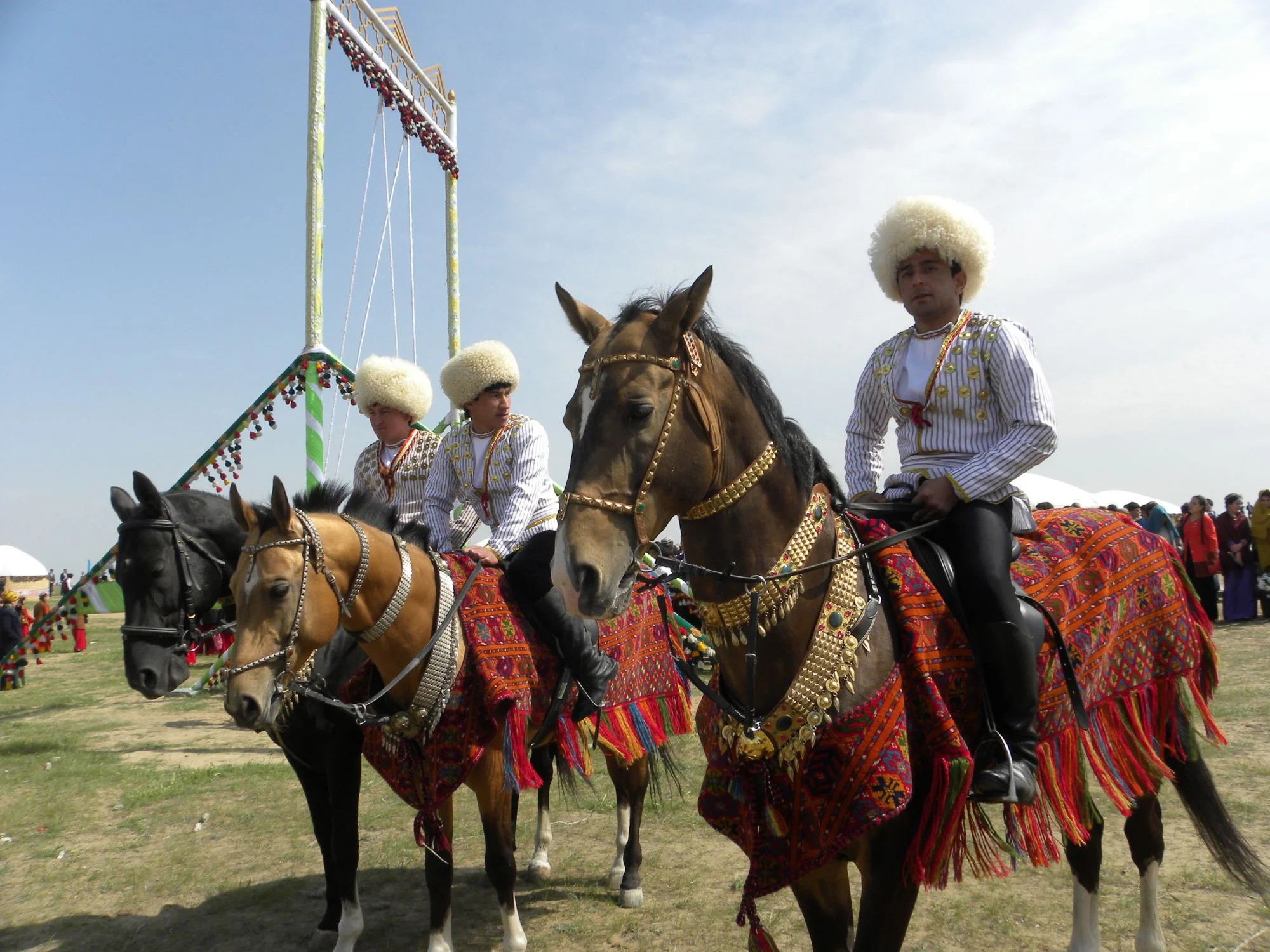 Riders in white sheepskin hats on Akhal-Teke horses in ornate carpet saddlecloths at a festival